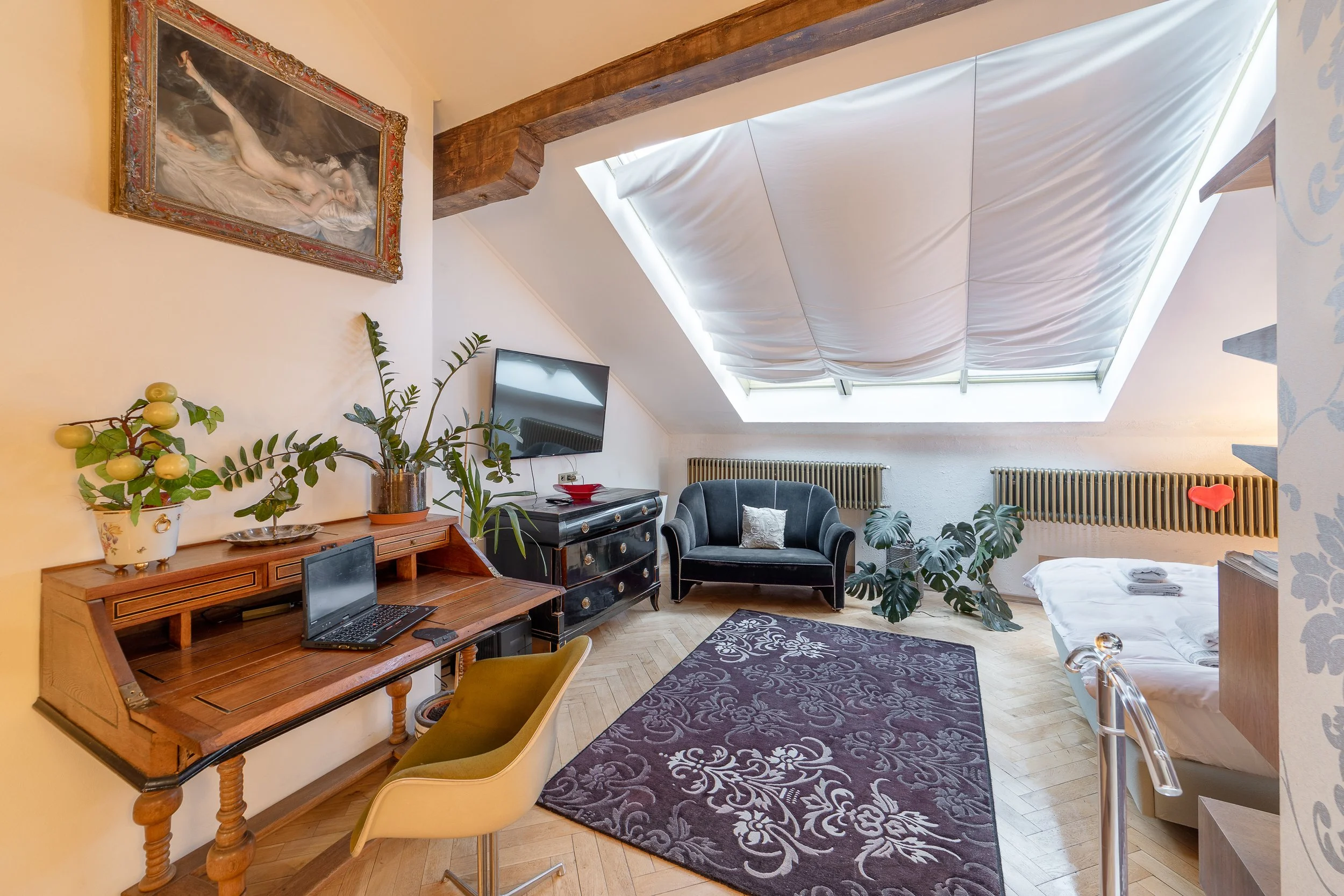 A cozy attic space with a pitched ceiling featuring a large skylight draped with white fabric. The room is decorated with plants, a vintage desk with a laptop, a black armchair, a bed with neatly folded towels, a flat-screen TV, a framed painting of 