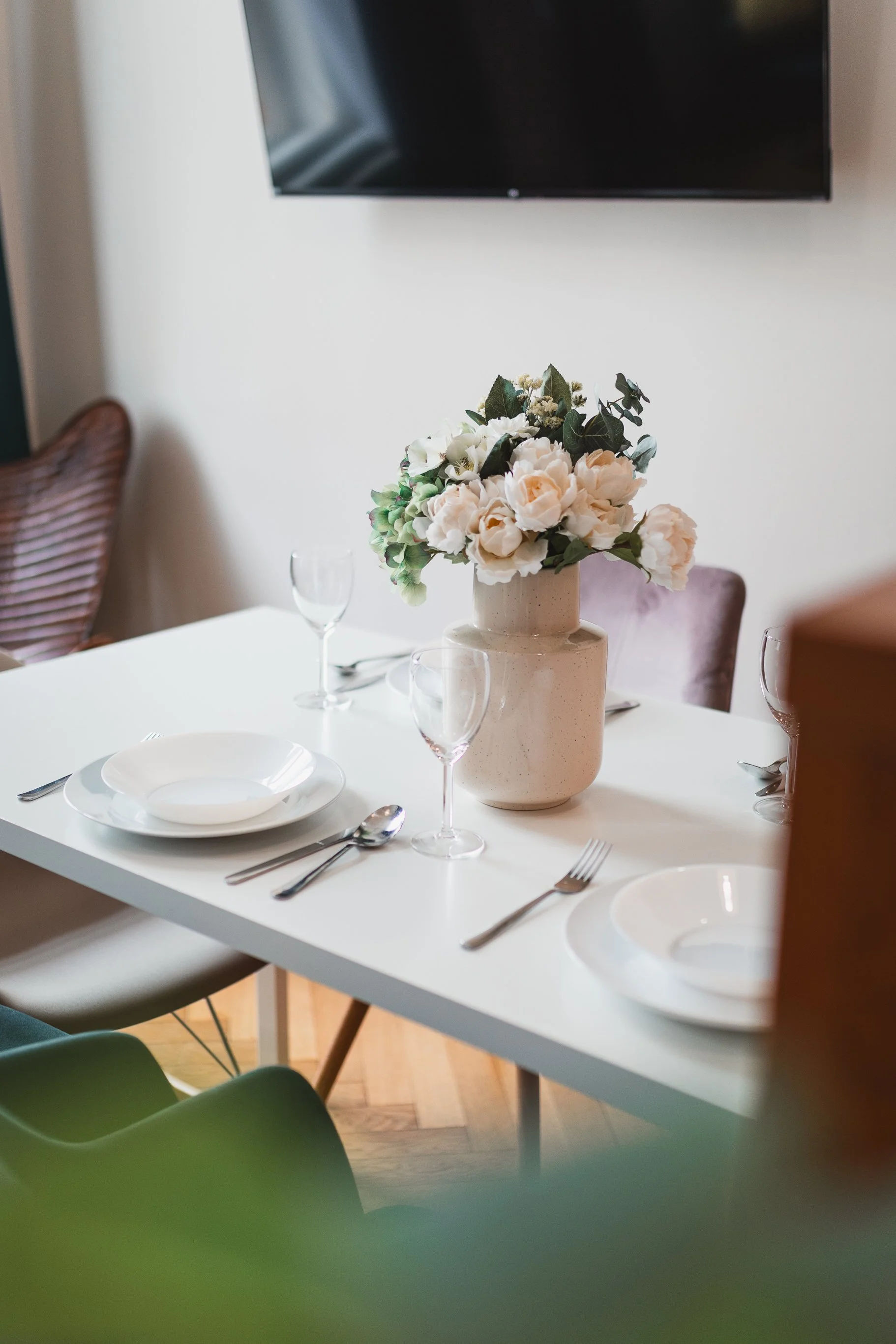 A dining table set with white plates, silverware, and glasses. In the center, a beige vase holds a bouquet of white and light pink flowers with green leaves. A television is mounted on the wall above the table.