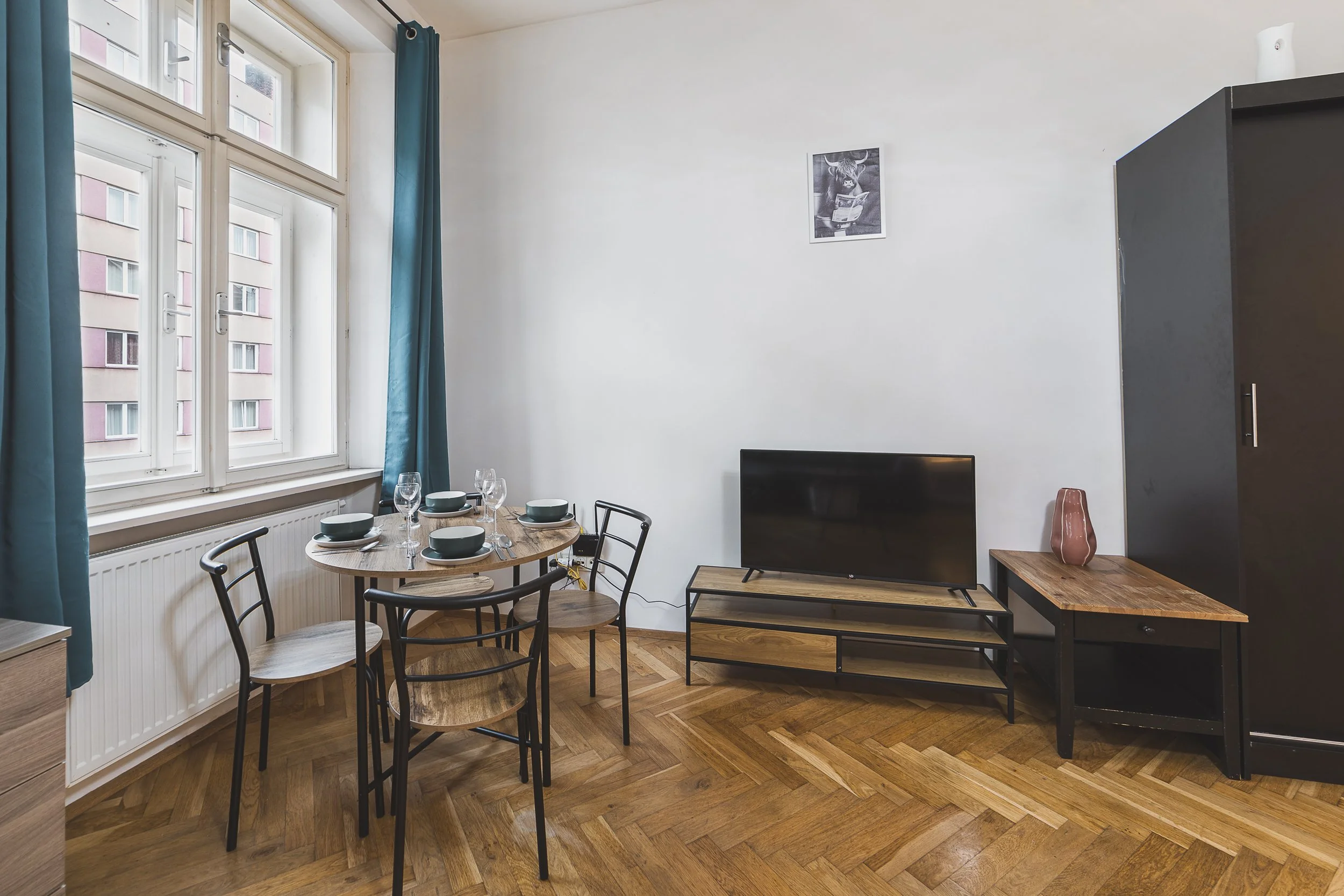 A dining area with a round wooden table set with four wine glasses and bowls, surrounded by black metal chairs, near large windows with blue curtains, a wall-mounted TV on a wooden stand, a tall dark gray wardrobe, a wooden side table with a pink vas