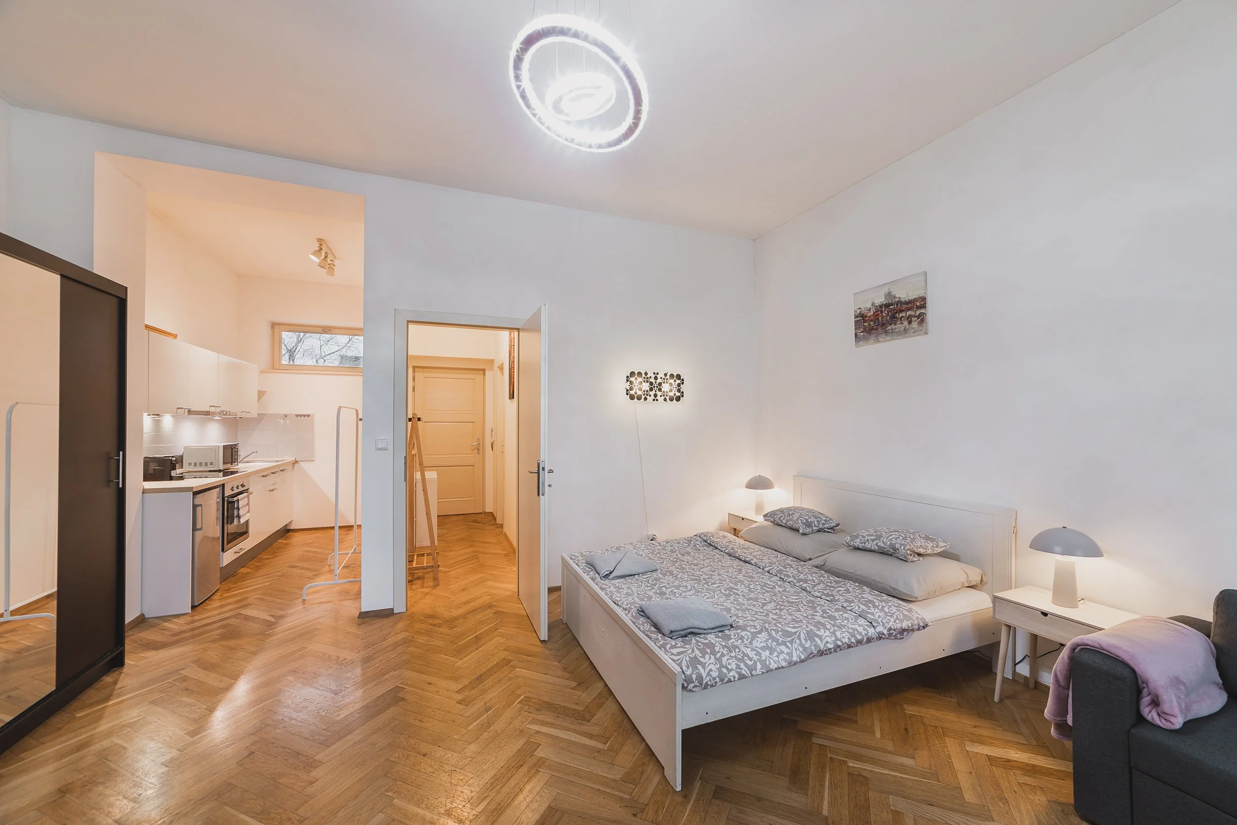 A cozy bedroom with a white bed, bedside lamps, and a small art picture on the wall. The room has hardwood floors and an open kitchen area visible in the background.