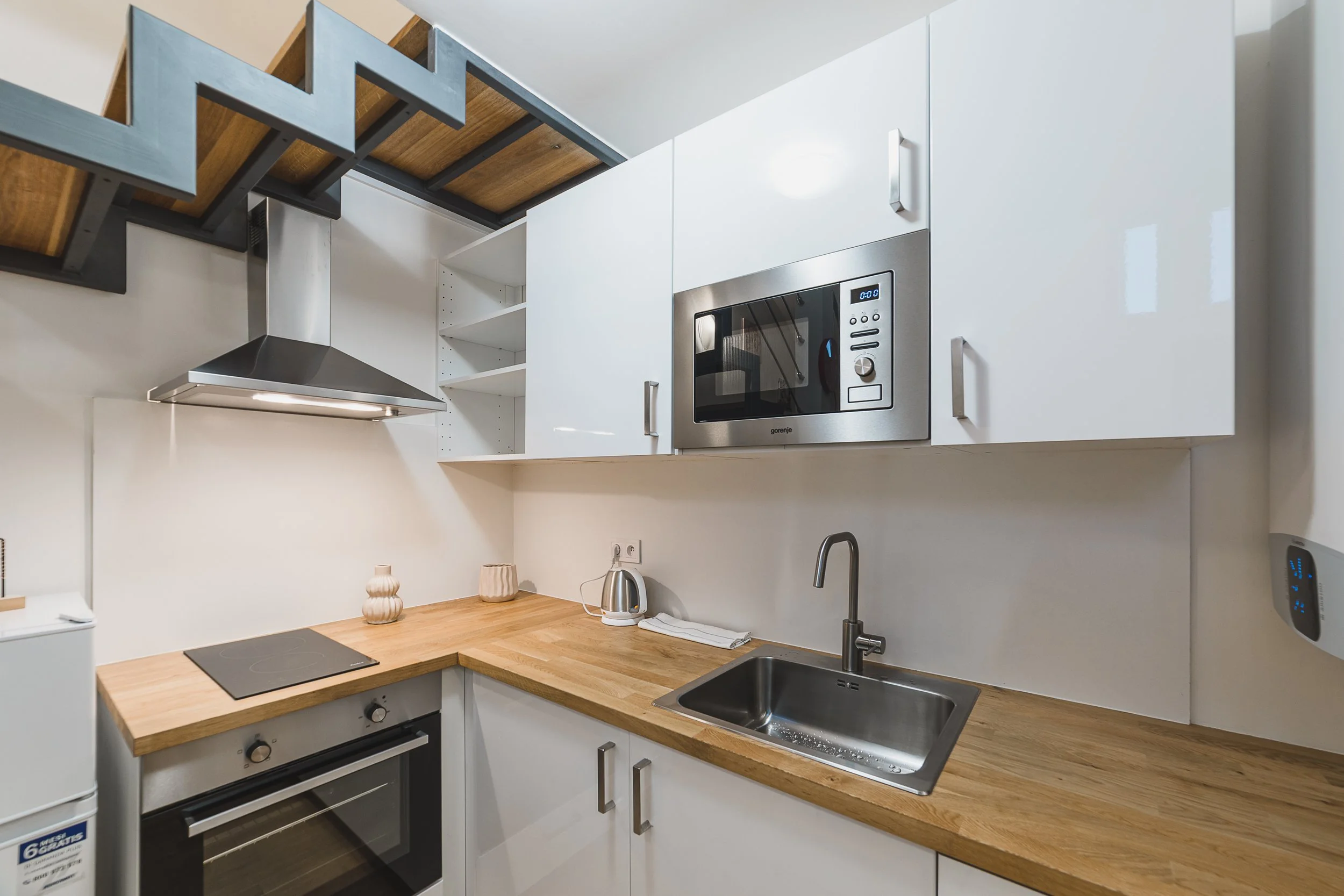Modern kitchen with white cabinets, wooden countertops, stainless steel microwave and range hood, black electric cooktop, stainless steel sink, and small decorative vases on the counter.