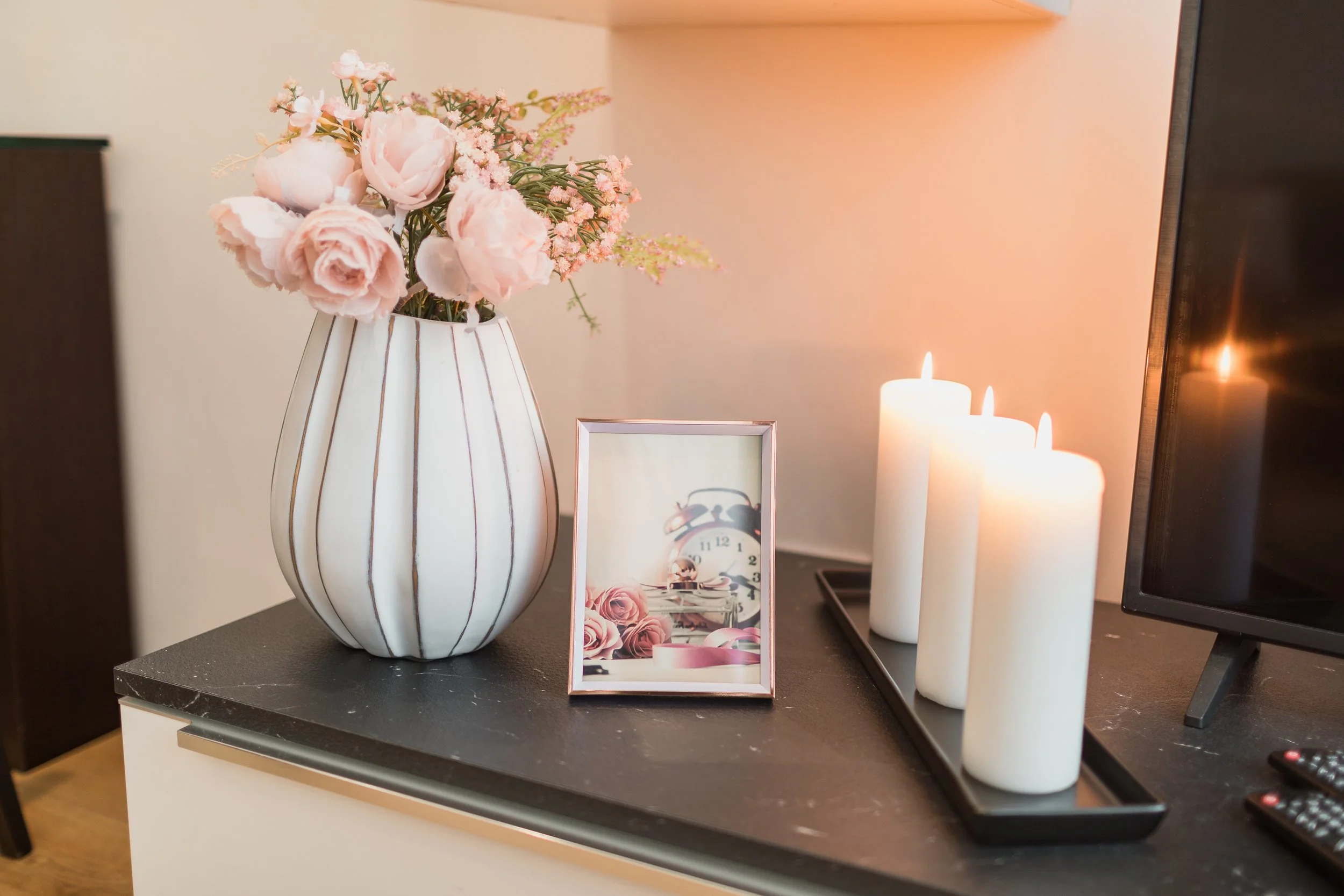 Pink and white artificial flowers in a white striped vase placed on a dark table, with candles and a framed picture nearby.