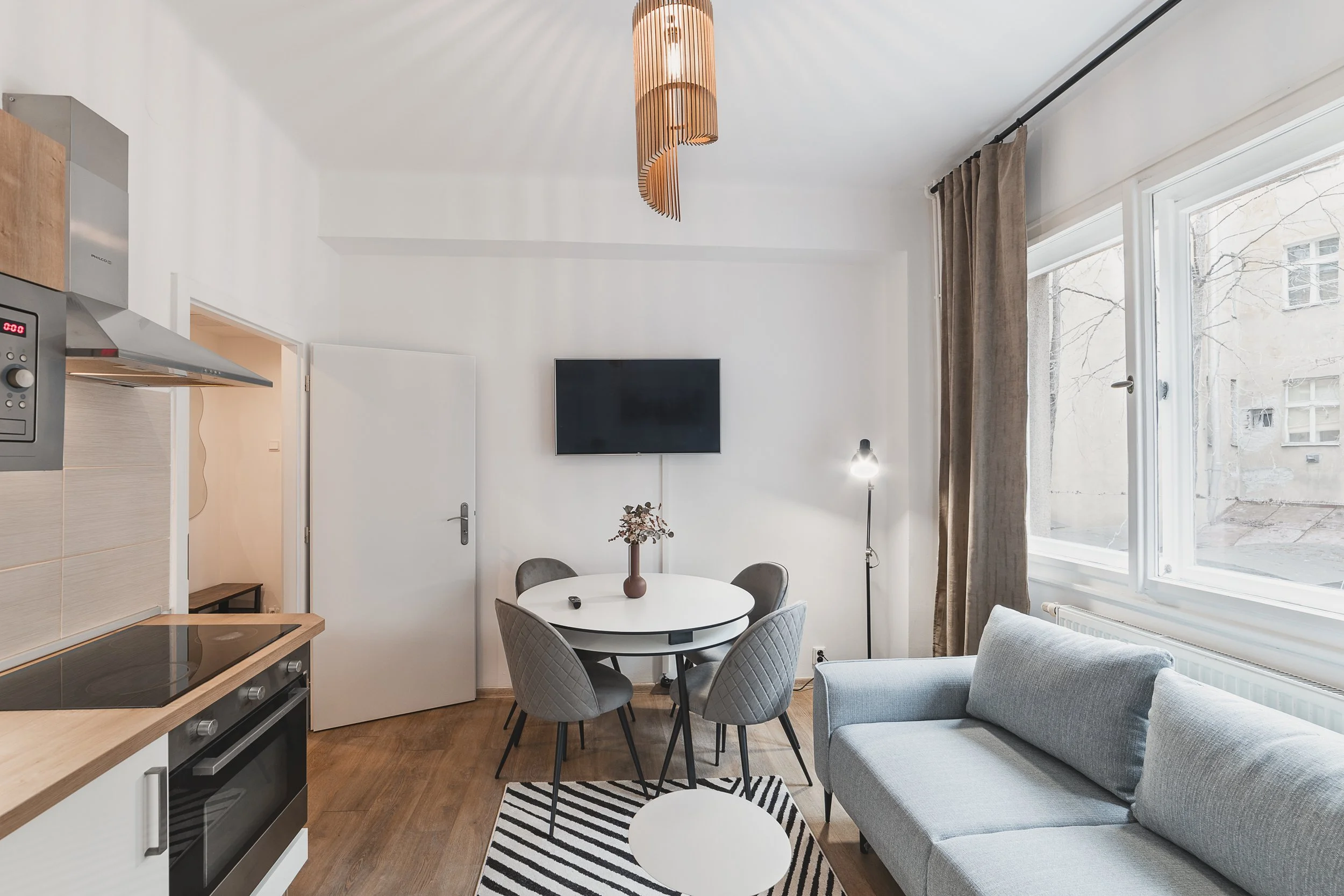 A modern kitchen and dining room with white walls, a round dining table with four gray quilted chairs, a gray sofa, a wall-mounted flat-screen TV, large windows with beige curtains, a contemporary wooden pendant light, and a striped rug on wooden flo
