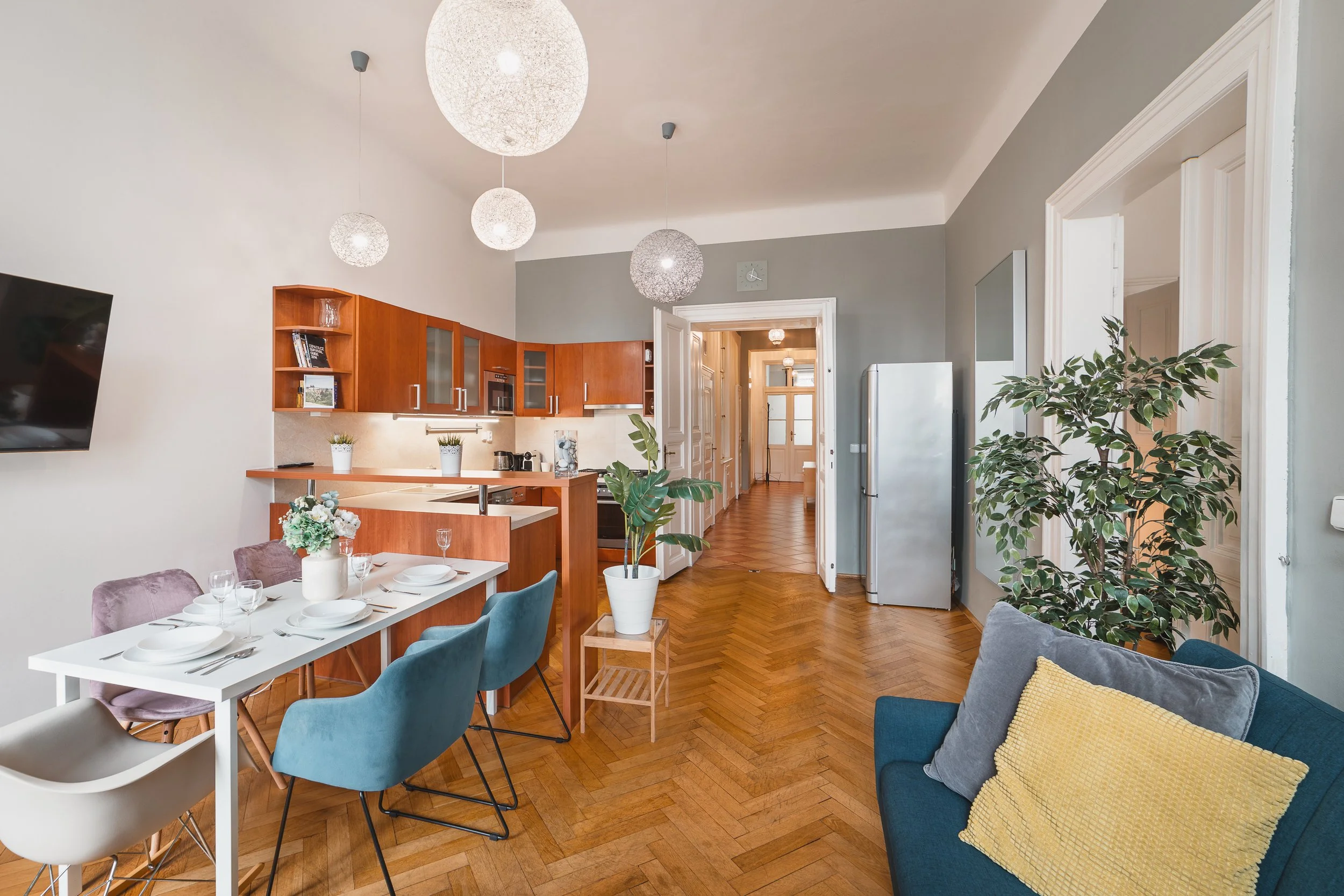 A modern living room with a dining area, eco-friendly pendant lights, a wooden kitchen cabinet, and several large potted plants, hardwood floors, and a mix of colorful chairs around a white dining table.