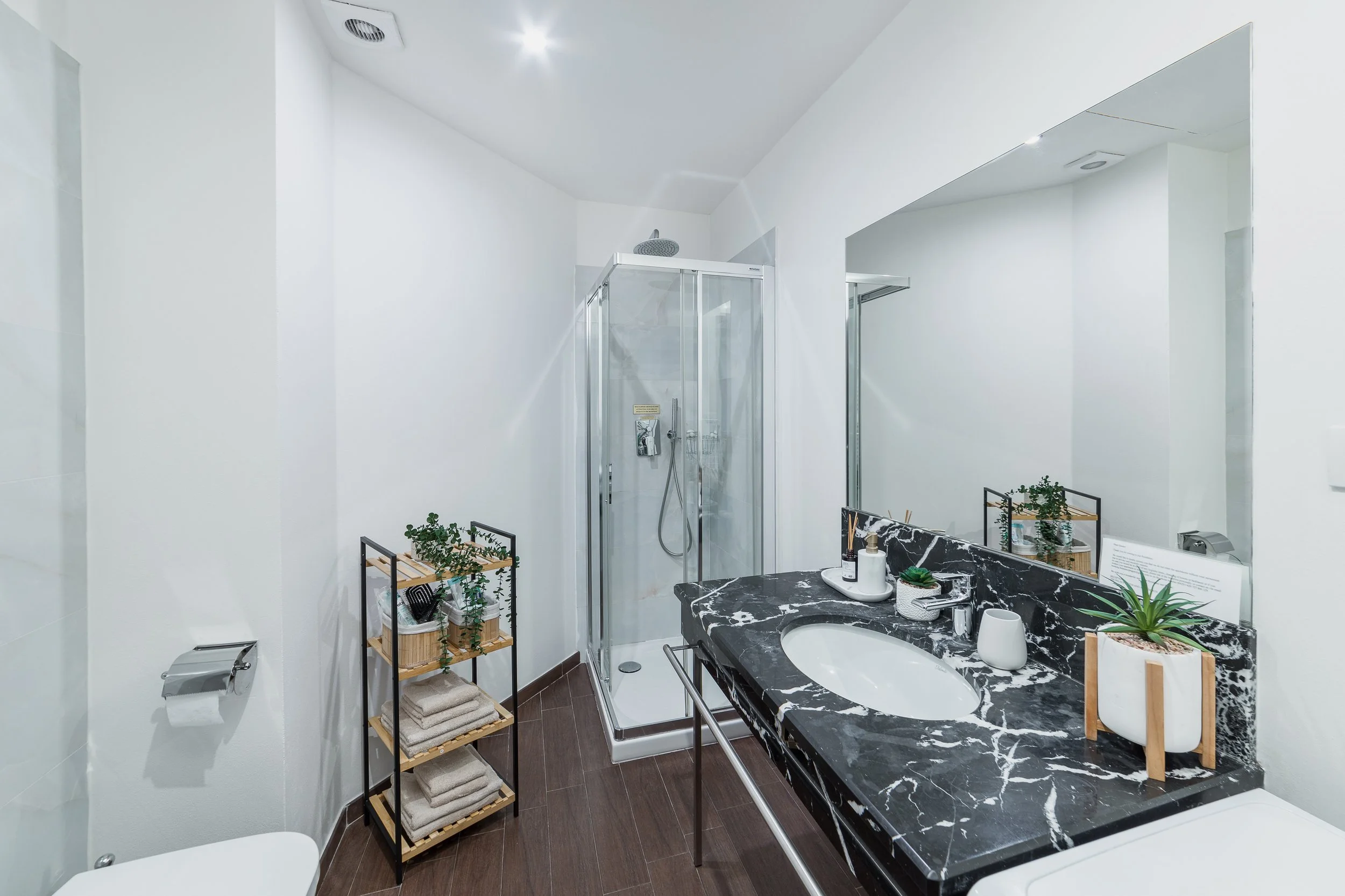 A modern bathroom with white walls, a glass shower enclosure, a black marble countertop sink, and a small shelving unit with towels and plants.