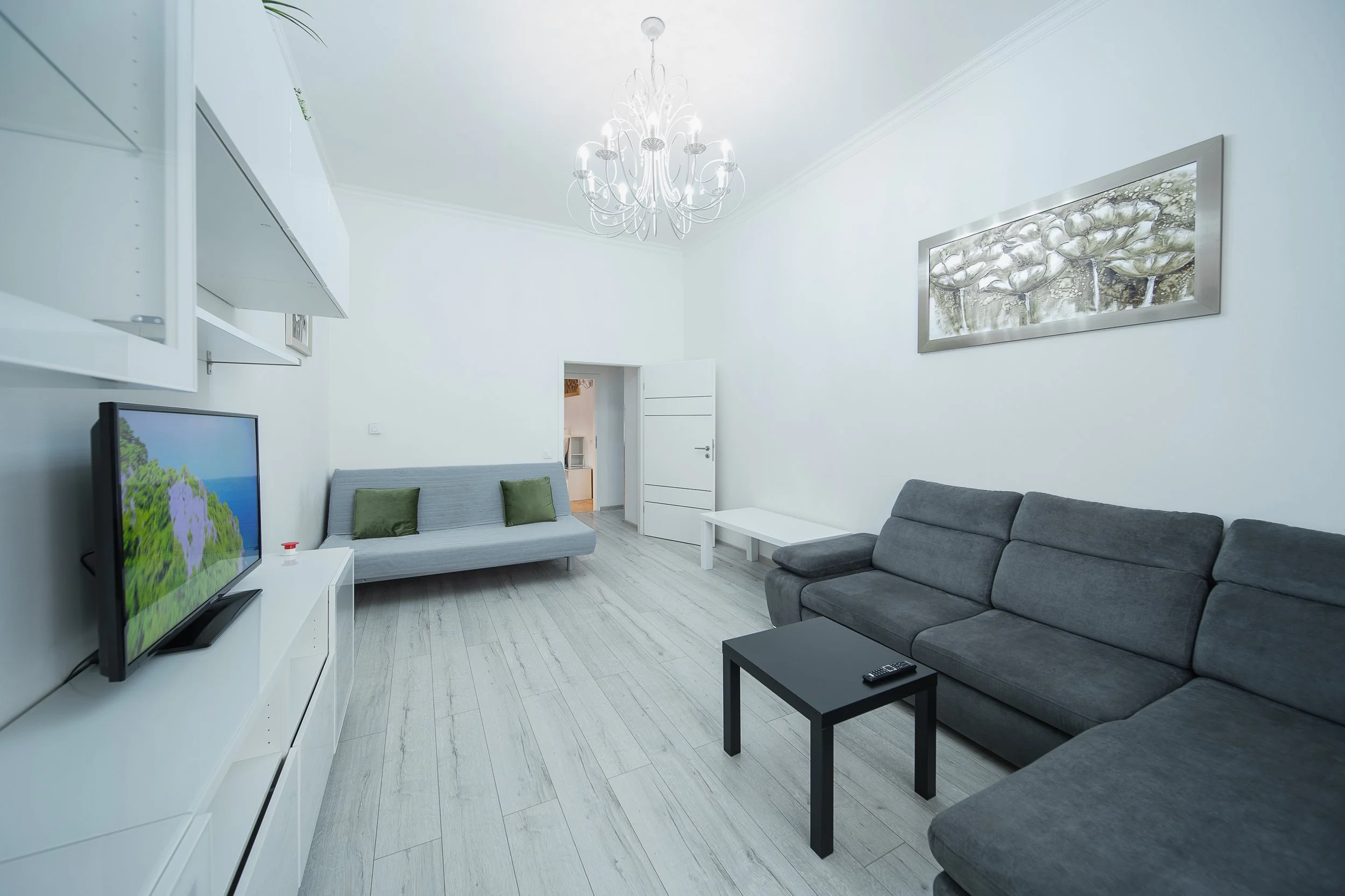 A modern living room with white walls, a chandelier, a gray sofa with green pillows, a white TV stand with a flat-screen TV, a small black coffee table with a remote, and artwork of flowers on the wall.