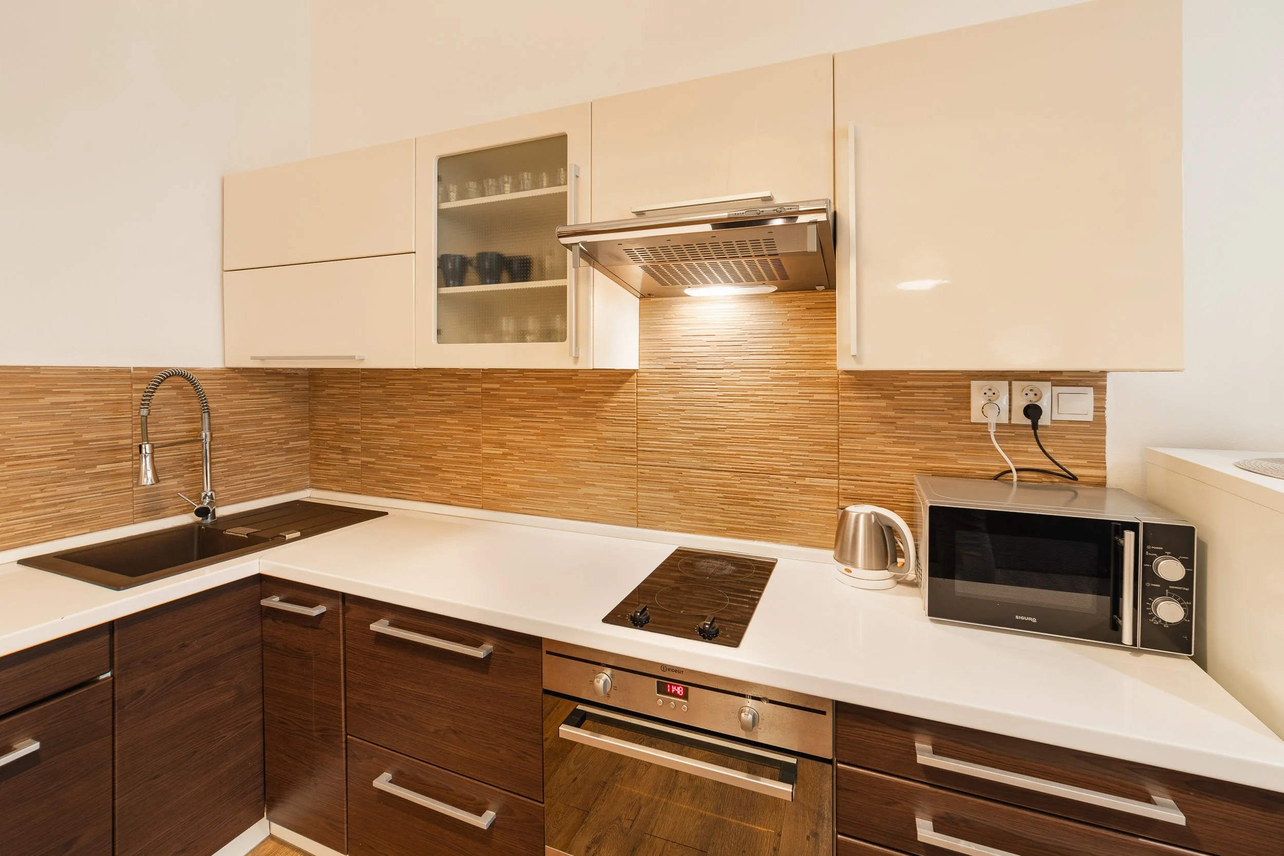 Modern kitchen with beige upper cabinets and dark wood lower cabinets, a backsplash with horizontal wooden slats, a stainless steel sink with a gooseneck faucet, a microwave oven on the right, a built-in oven below, and electrical outlets on the wall