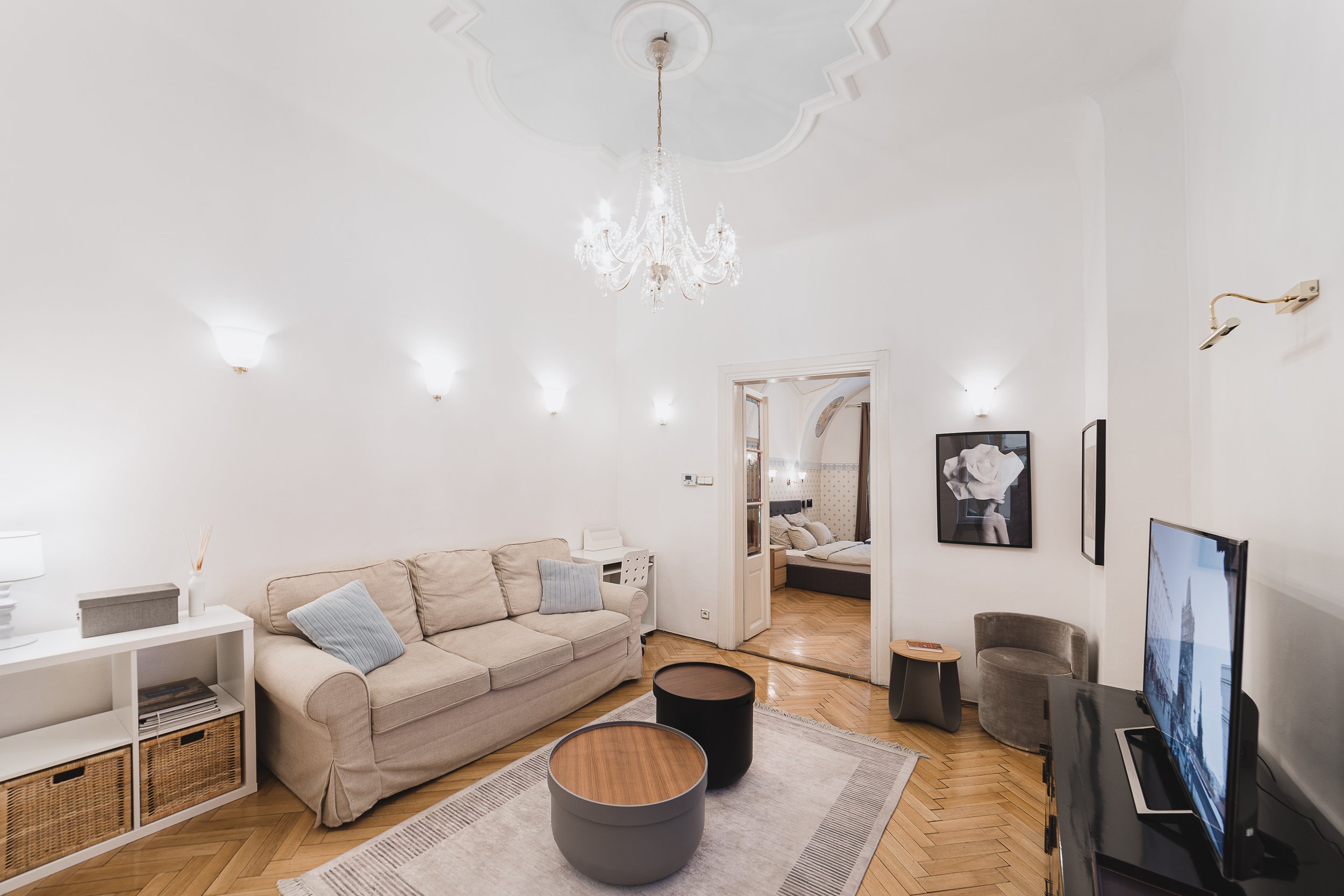 Living room with beige sofa, two round coffee tables, and a TV. Bright white walls with wall sconces, a chandelier, and a painting. Doorway leads to a bedroom with a bed and patterned wallpaper.