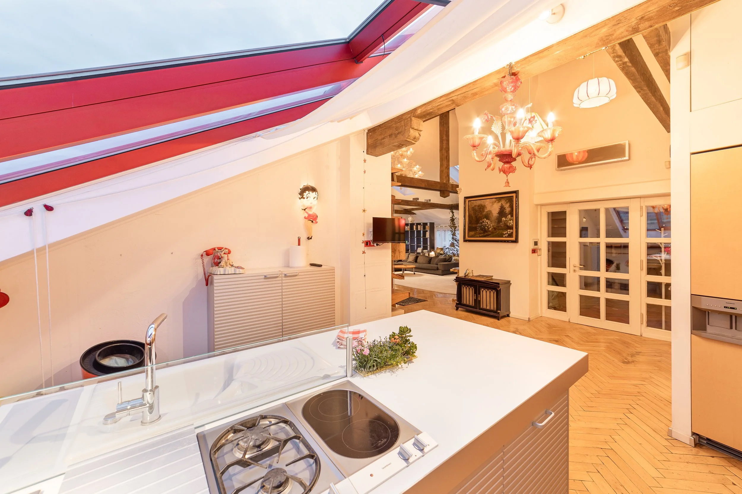 Interior view of a living space with a kitchen in the foreground and a living room in the background. The kitchen has a white countertop with a built-in sink and electric stove. The ceiling has exposed wooden beams, and a pink chandelier hangs from i