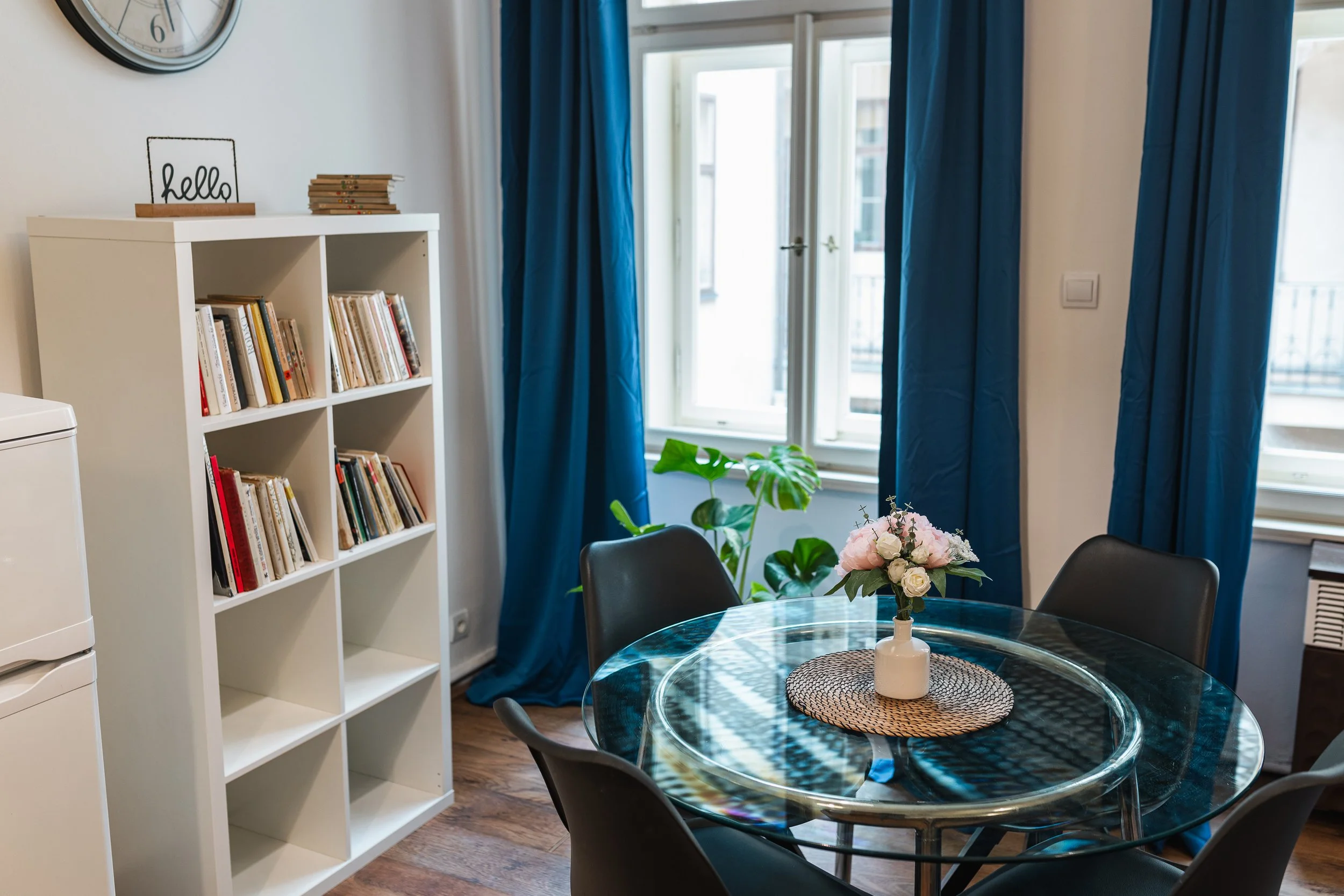 A cozy indoor space featuring a round glass dining table with a flower arrangement, black chairs, a white bookshelf filled with books, blue curtains over a large window, and a clock on the wall.