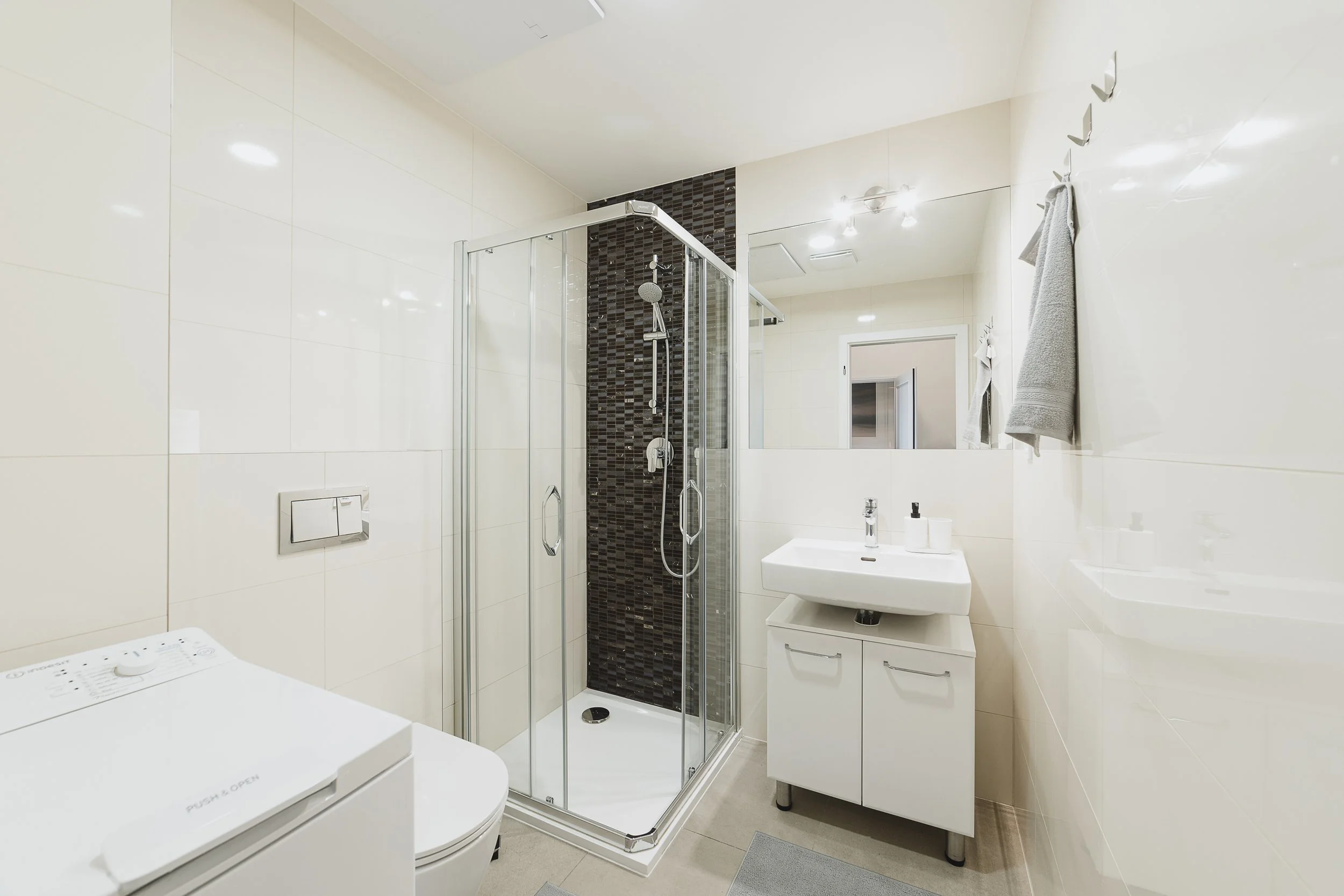 Modern bathroom with white walls, a shower stall with dark mosaic tiles, a small sink with cabinet, and a towel hanging on hooks on the wall.