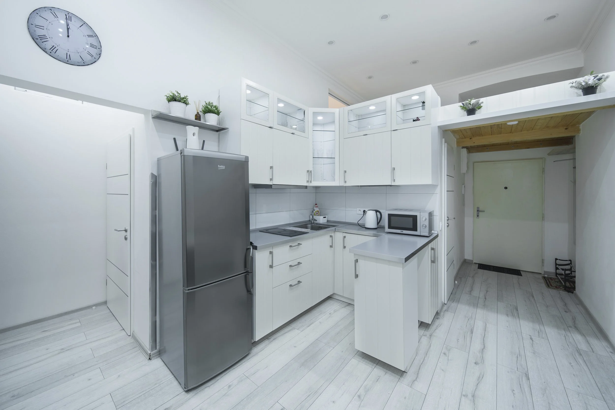 Modern white kitchen with cabinets and appliances, including a stainless steel refrigerator, microwave, electric cooktop, kettle, and decorative plants on upper shelves. There is a clock with Roman numerals on the wall and a door in the background.