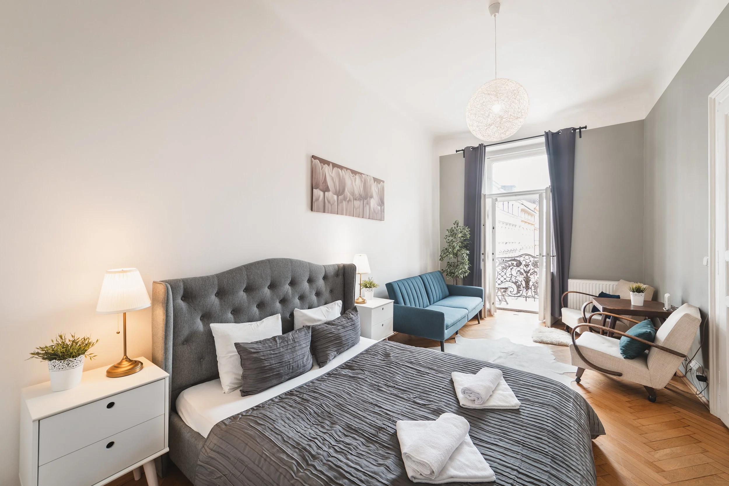 A bright, cozy living room with a large bed, grey headboard, white nightstand with a plant and lamp, a blue sofa, and a white armchair with wooden arms near the window. There are curtains, a framed flower painting, and a balcony door with iron railin