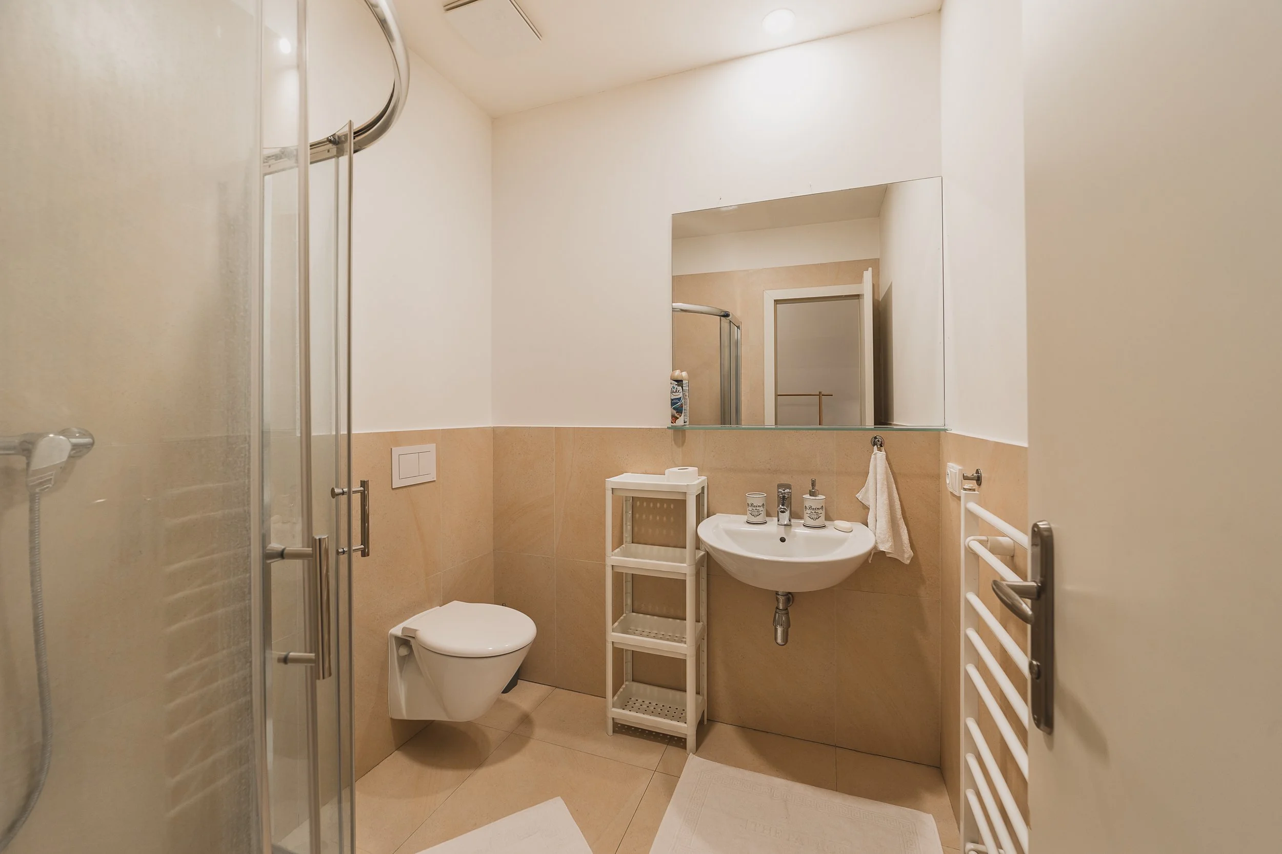 A compact bathroom with beige tiled walls and a white ceiling. It features a small white sink with a mirror above, a handheld shower with frosted glass door, a white toilet, and a white towel on a wall-mounted towel rack. A small white shelving unit 