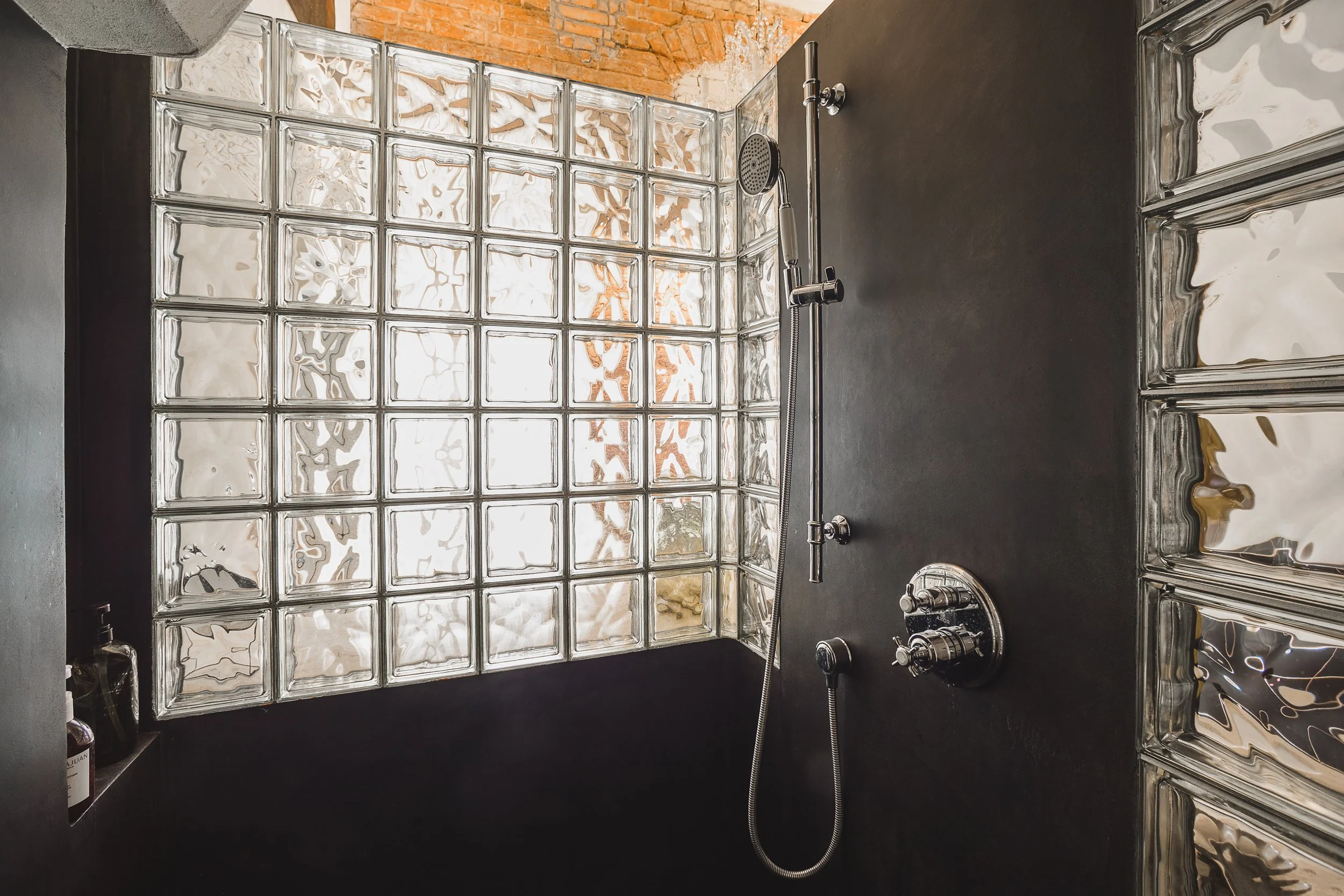 A modern shower area featuring a glass block wall, black walls, a rain shower head, and built-in shelves with bottles.
