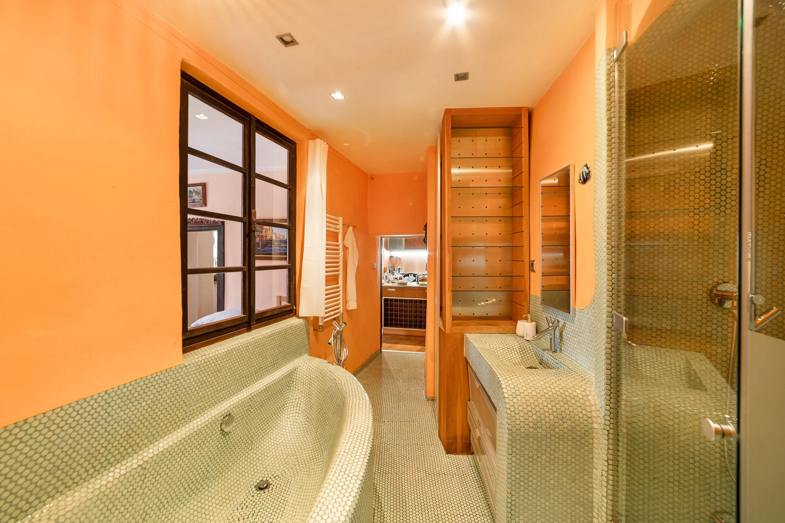 A bathroom with orange walls, a large window with dark wooden frames, and green mosaic tile on the bathtub, sinks, and floor. There is a wooden built-in shelving unit and a mirror above the sink. Towels are hanging on a towel rack and kitchen utensil