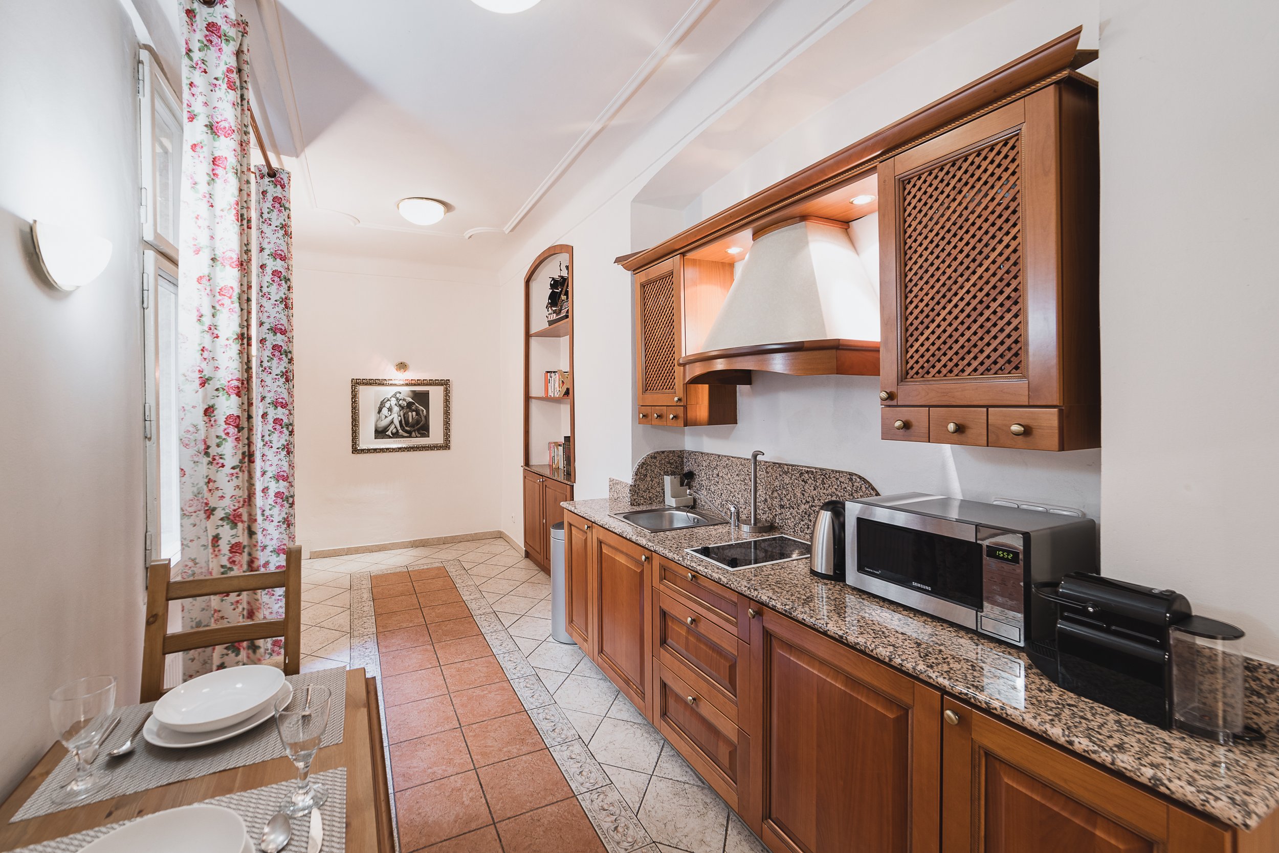 Kitchen with wooden cabinets, granite countertop, microwave, sink, and small table with place settings, floral curtains, and a framed black-and-white photo on the wall.