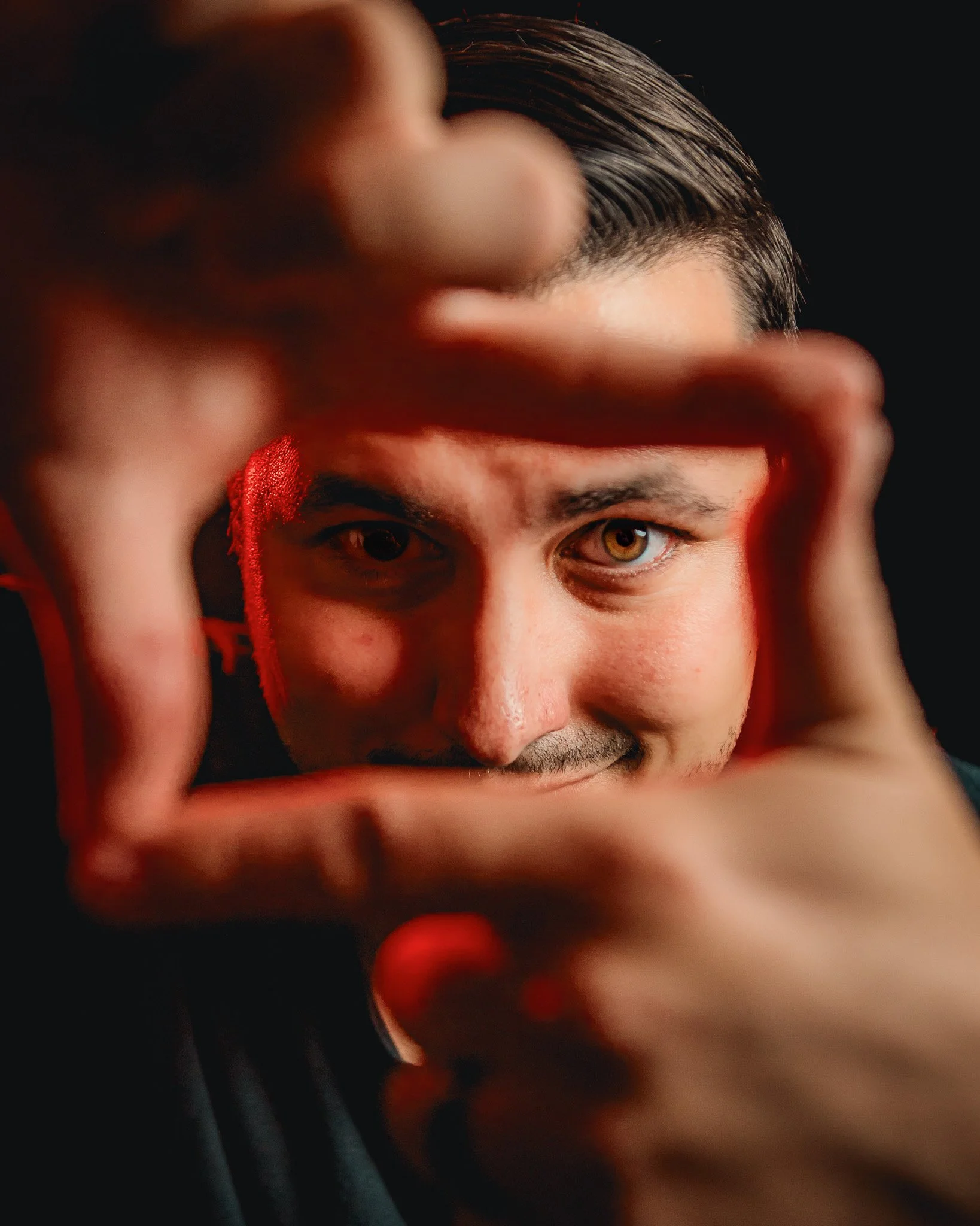 Close-up of a man's face with dark hair and brown eyes, framing his eyes with his hands against a dark background.