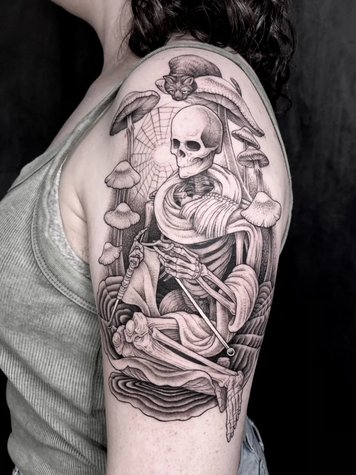 Fine line black and grey skeleton knitting a scarf and nestled amongst mushrooms, with a cat perched above, created by Seattle tattoo artist Eli Bischof.