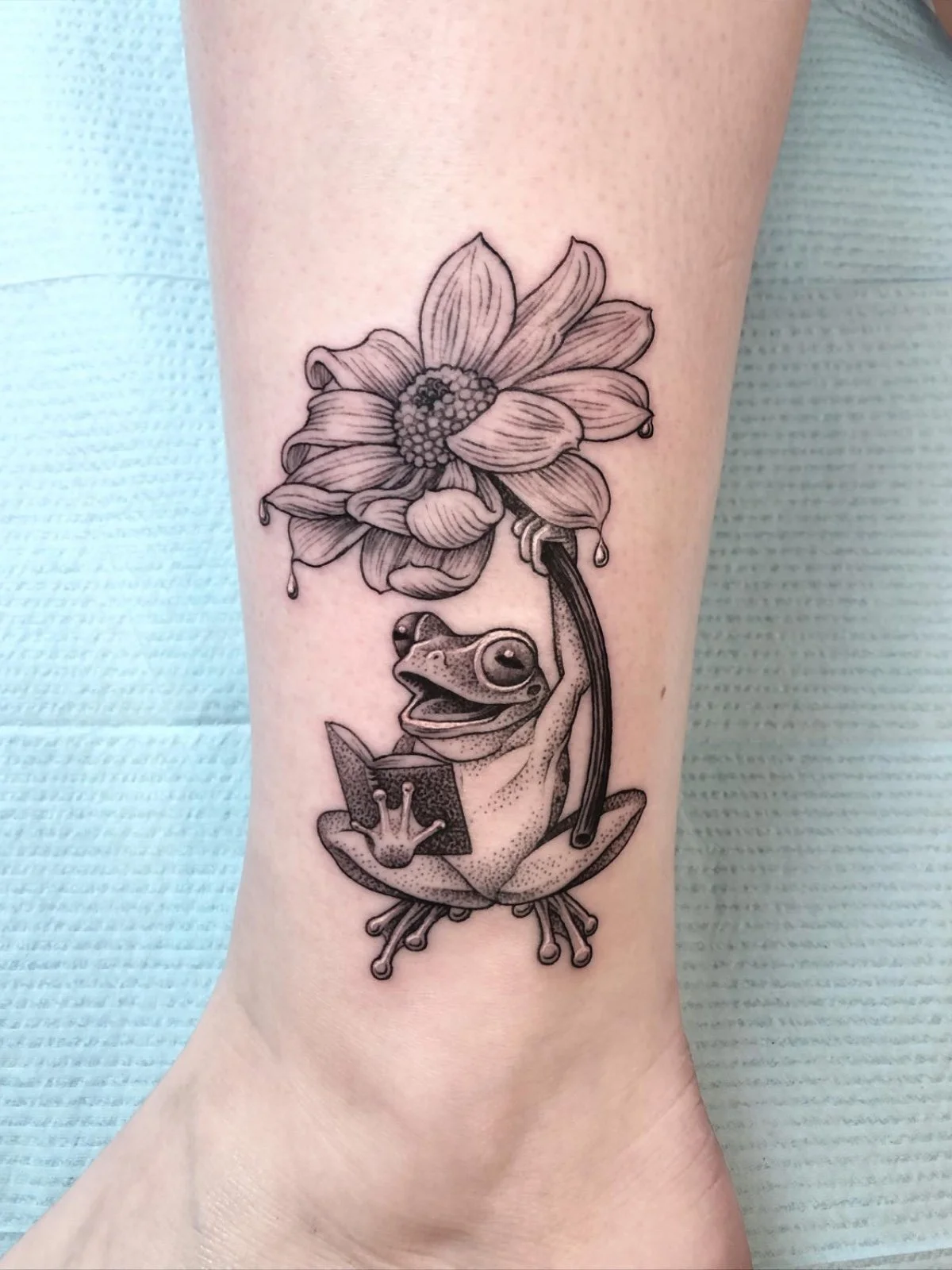 A fine line and black and grey tattoo of a cute frog holding a flower and reading a book, created by Seattle tattoo artist Eli Bischof.