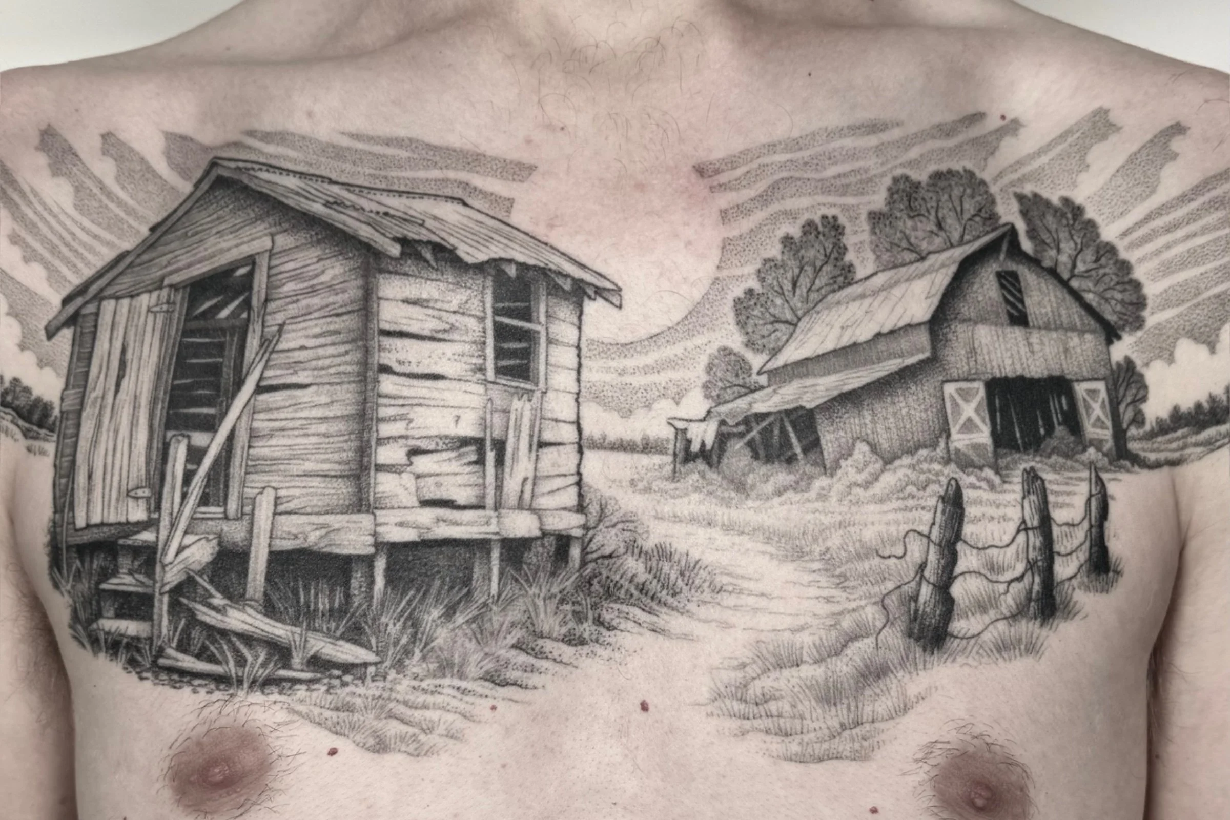 Healed illustrative black and grey chest tattoo by Seattle tattoo artist Eli Bischof featuring a weathered wooden shack, rustic barn, country road, and dramatic sky.