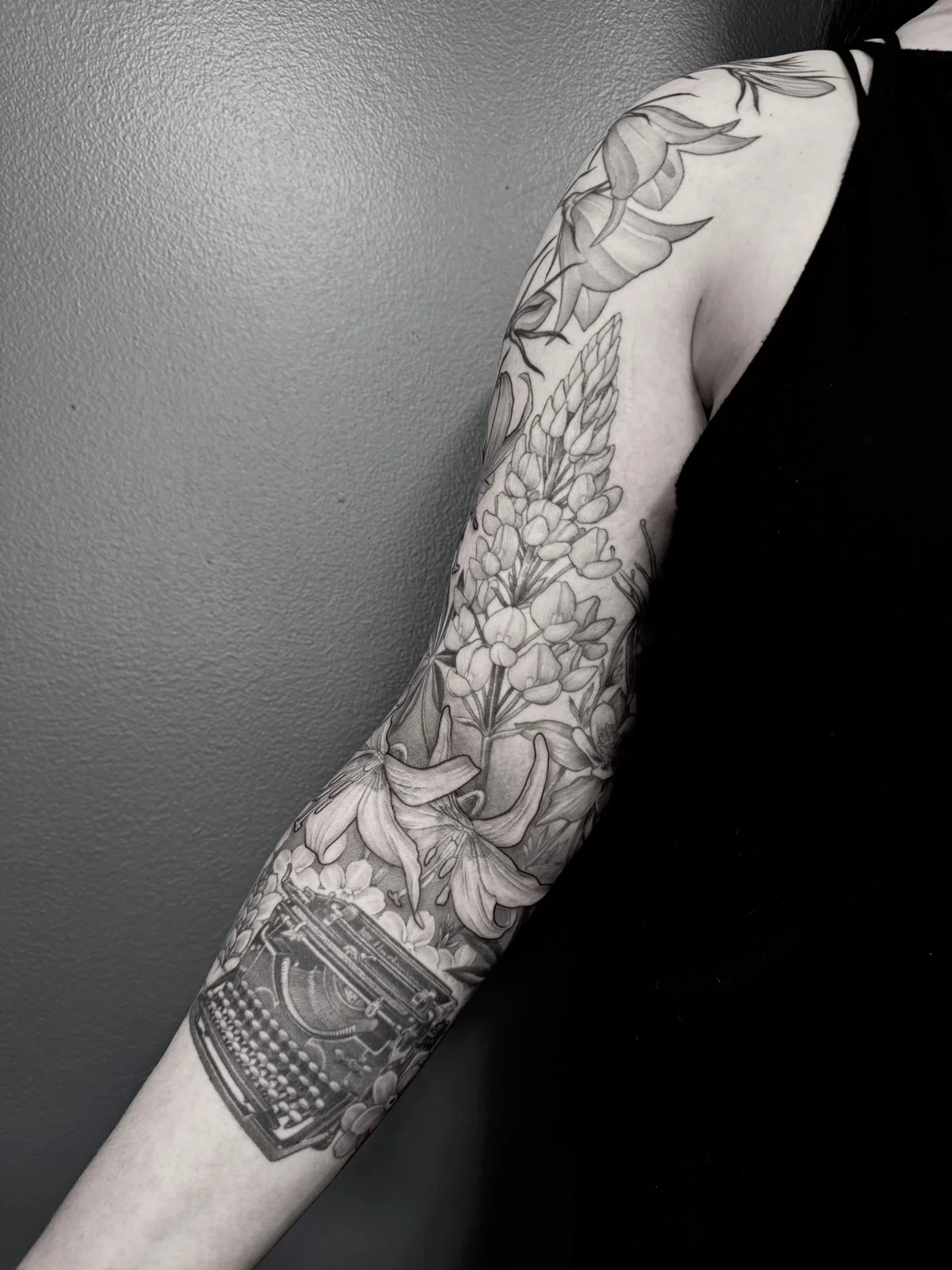 Fine line black and grey sleeve tattoo, with flowing flower arrangement and antique typewriter nestled amongst forget me not flowers, created by Seattle tattoo artist Eli Bischof