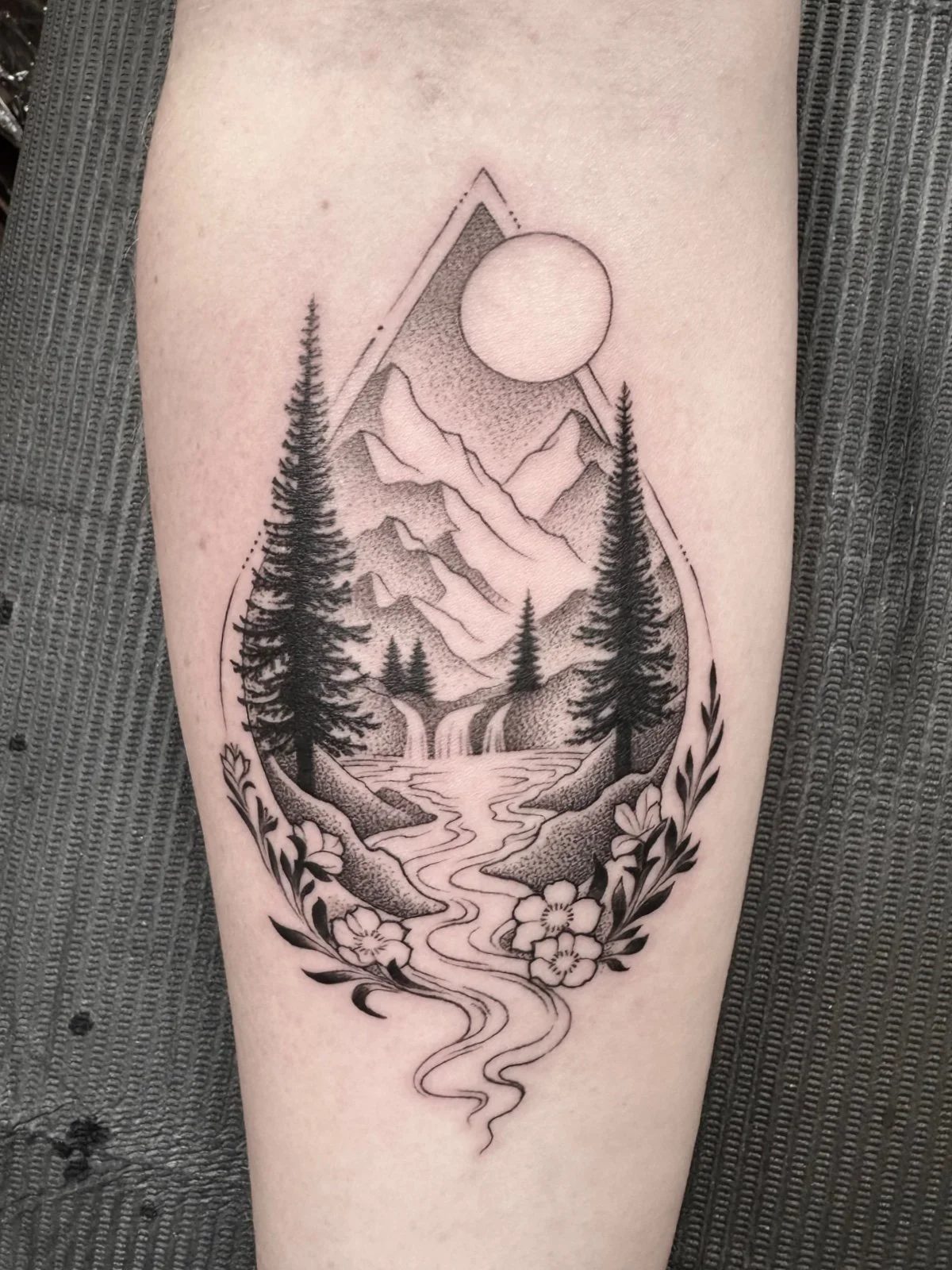 A delicate fine line black and grey landscape tattoo in the shape of a water drop, with flowers and leaves, created by Seattle tattoo artist Eli Bischof.