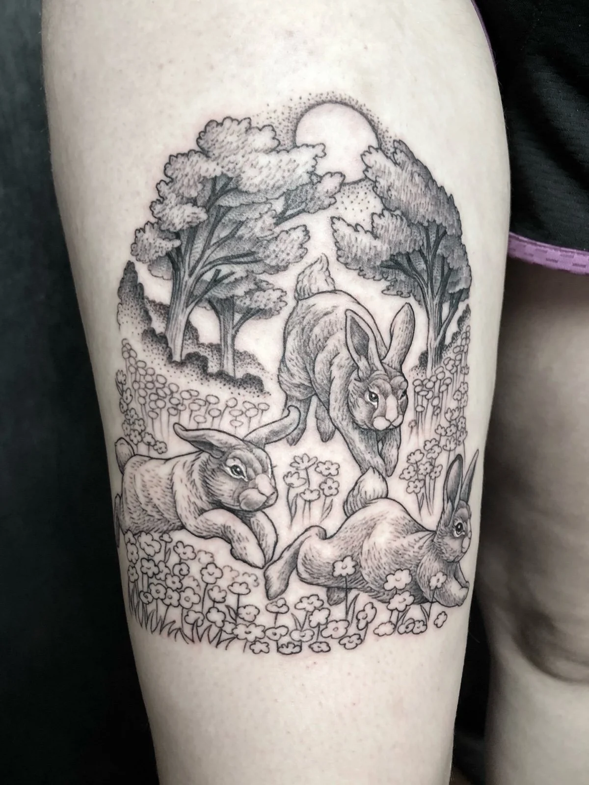 A fine line black and grey landscape tattoo, with three cottontail rabbits leaping through a flowery meadow and trees, created by Seattle tattoo artist Eli Bischof.