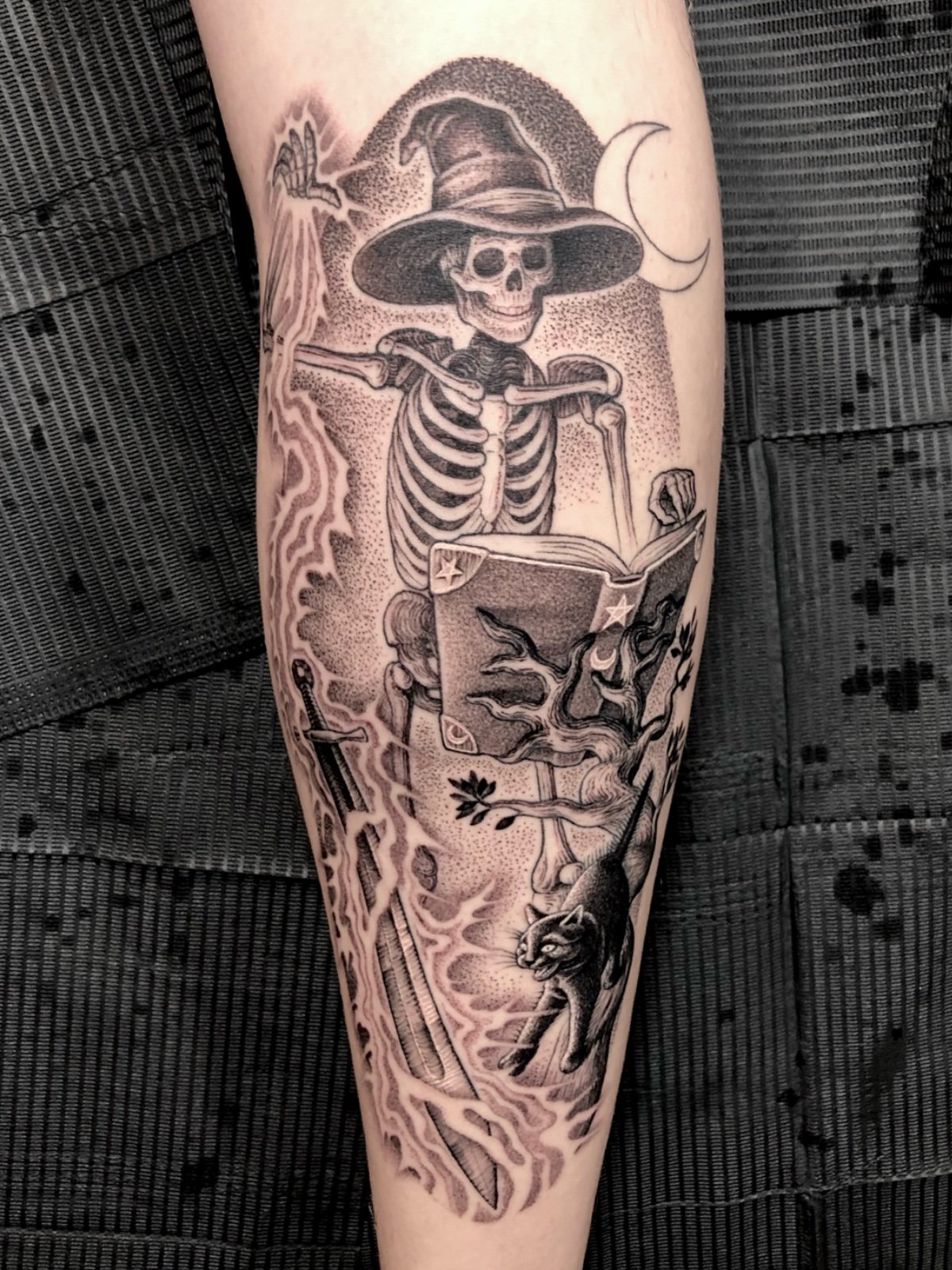 Fine line black and grey tattoo of a wizard skeleton summoning a sword, with a startled black cat, created by Seattle tattoo artist Eli Bischof.