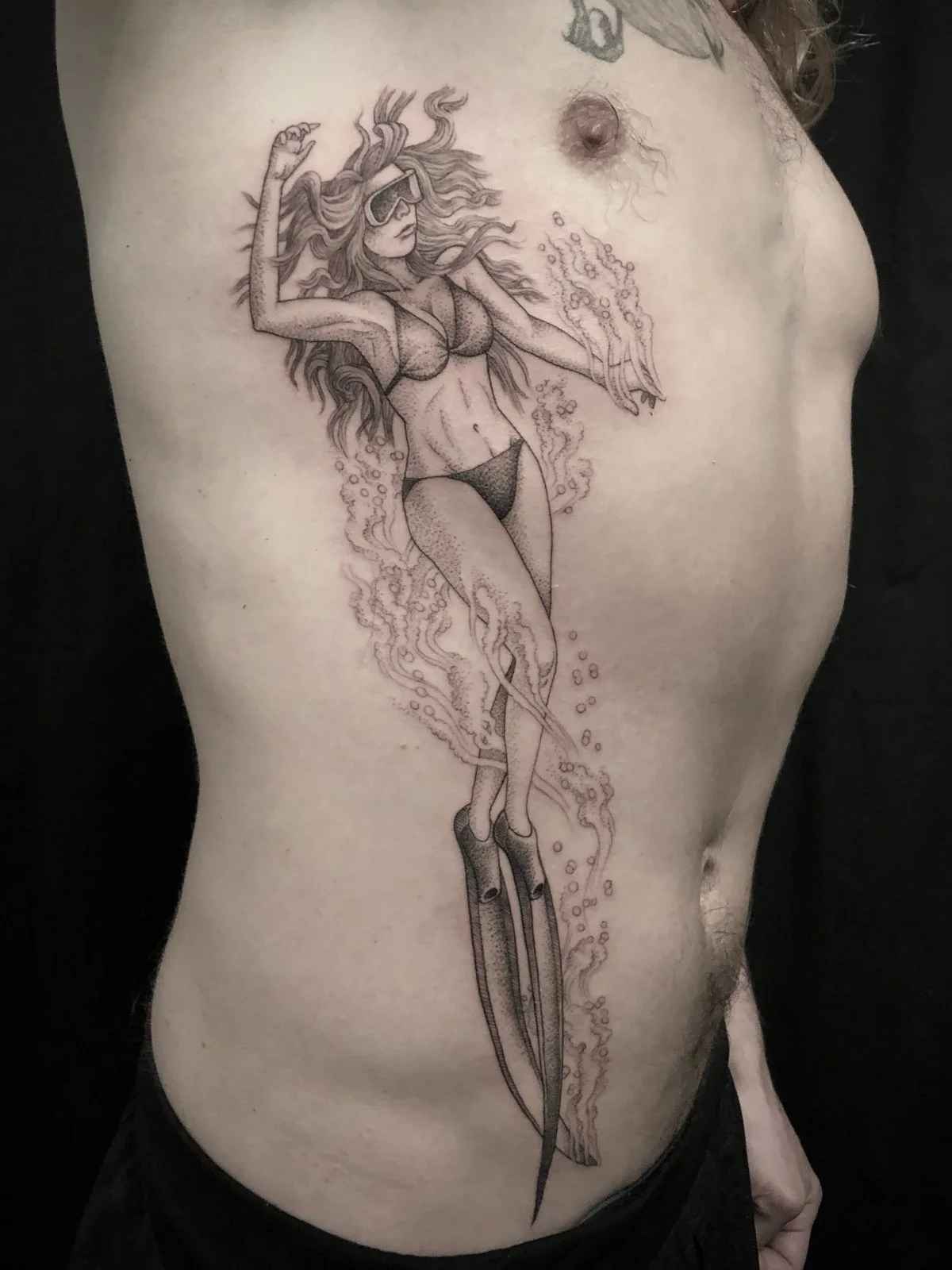 A fine line and black and grey tattoo with dotwork shading, depicting a young woman free diving on the torso, with bubbles coming from her flippers, created by Seattle tattoo artist Eli Bischof.