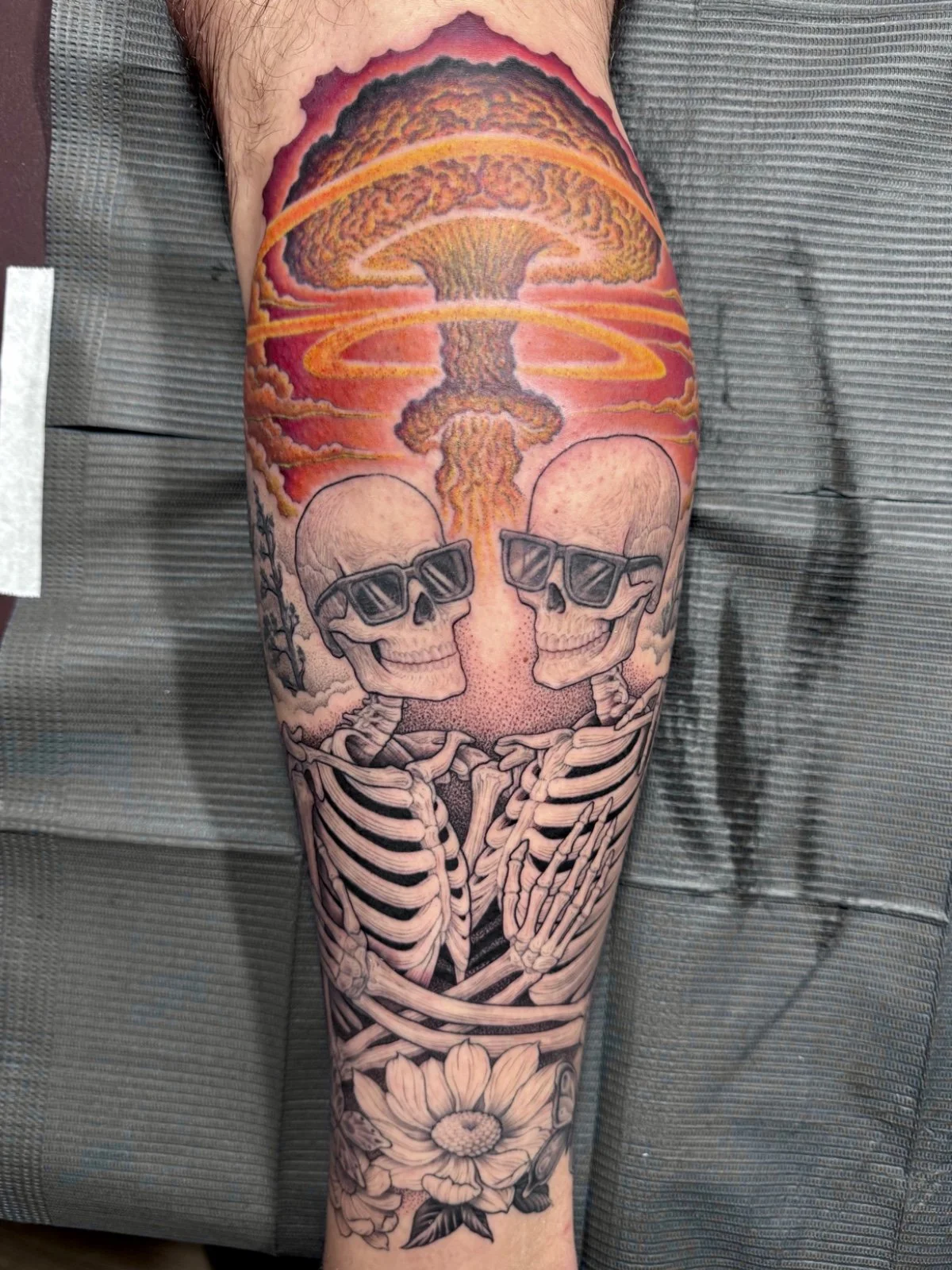 Fine line black and grey and color tattoo of two skeletons hugging and wearing sunglasses as a mushroom cloud rises behind them, created by Seattle tattoo artist Eli Bischof.