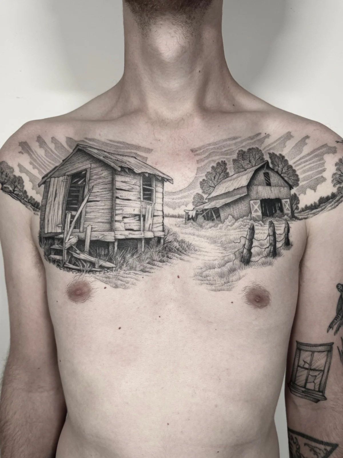 A fine line black and grey landscape tattoo spanning the chest, featuring an abandoned farmyard, created by Seattle tattoo artist Eli Bischof.