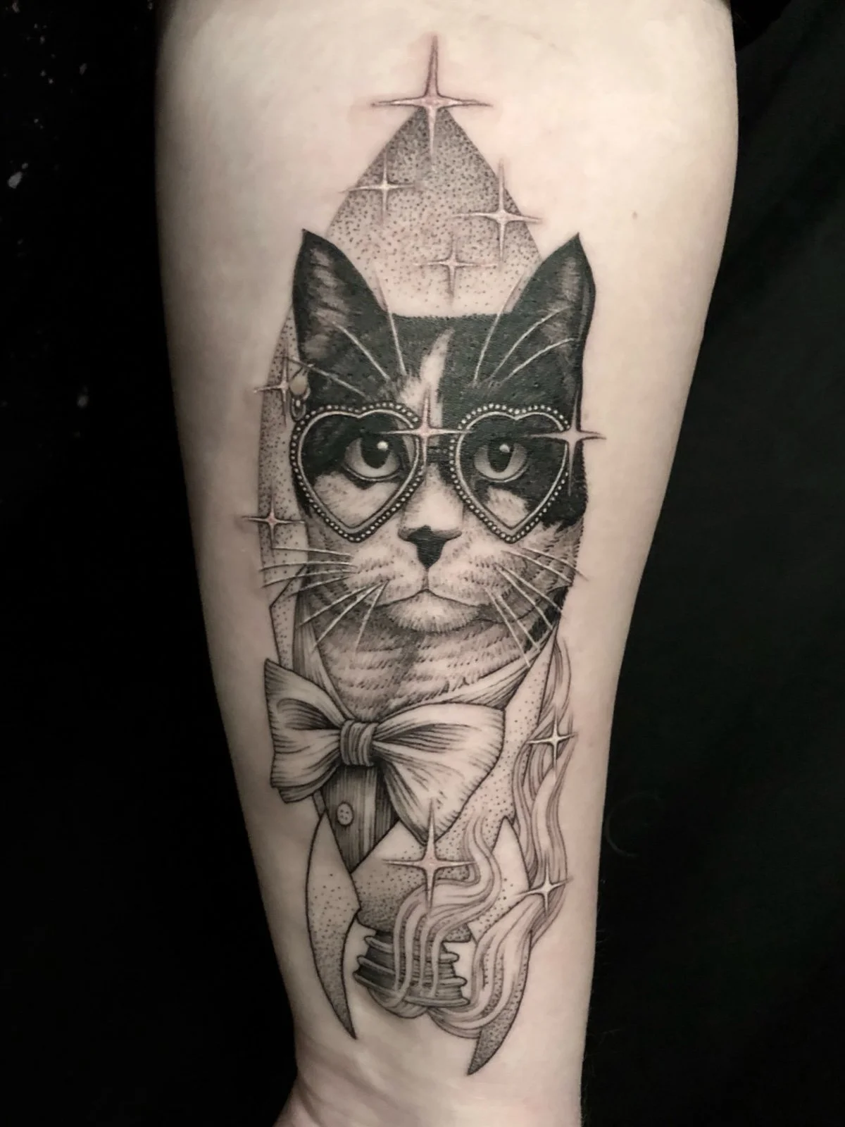 Fine line and black and grey whimsical pet portrait tattoo of a tuxedo cat wearing a bow tie and heart shaped glasses, coming out of a rocket shape in tribute to Elton John, created by Seattle tattoo artist Eli Bischof.