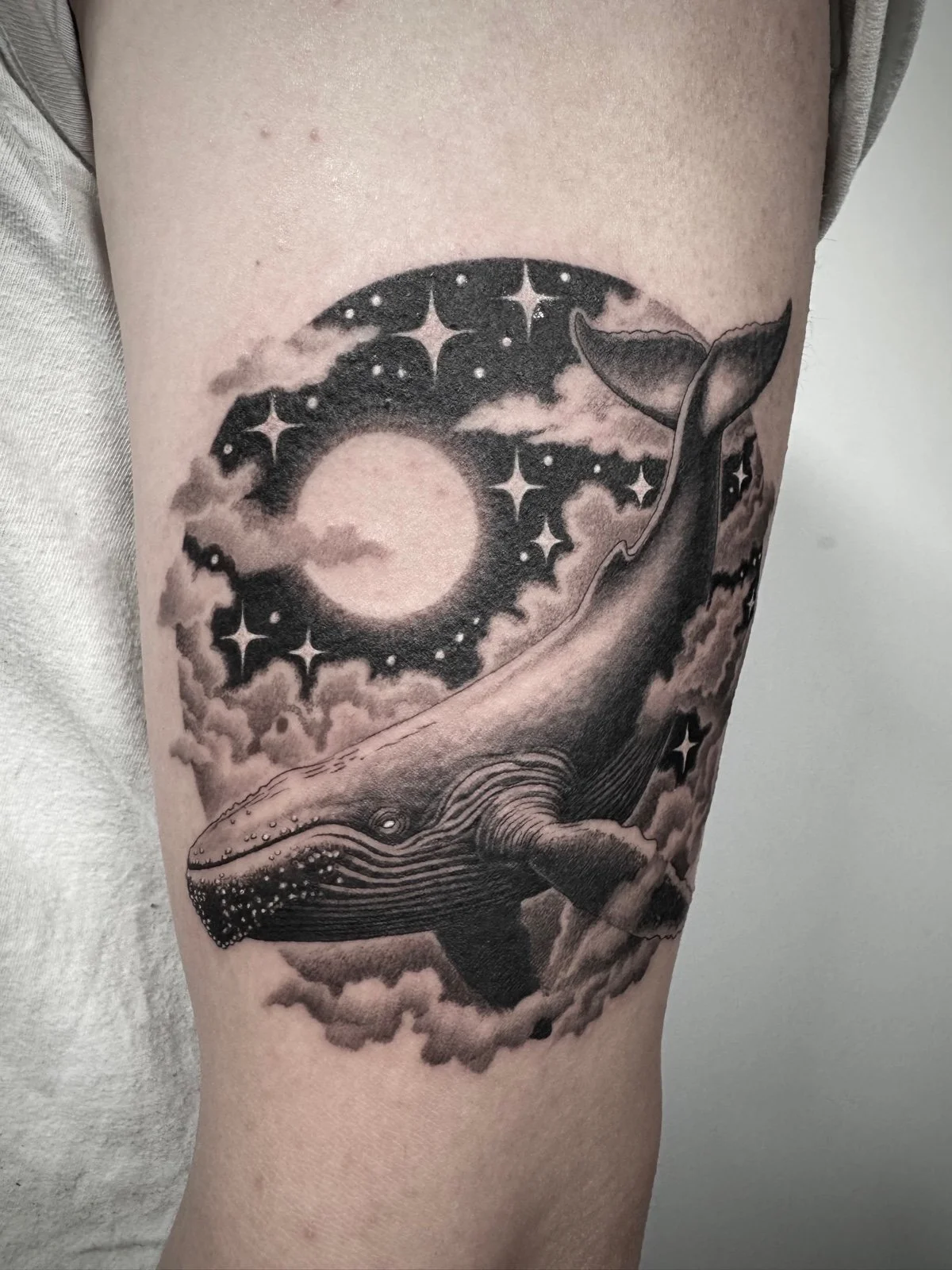 A fine line and black and grey tattoo of a surreal scene, with a humpback whale swimming through clouds under the moonlight and stars, created by Seattle tattoo artist Eli Bischof.