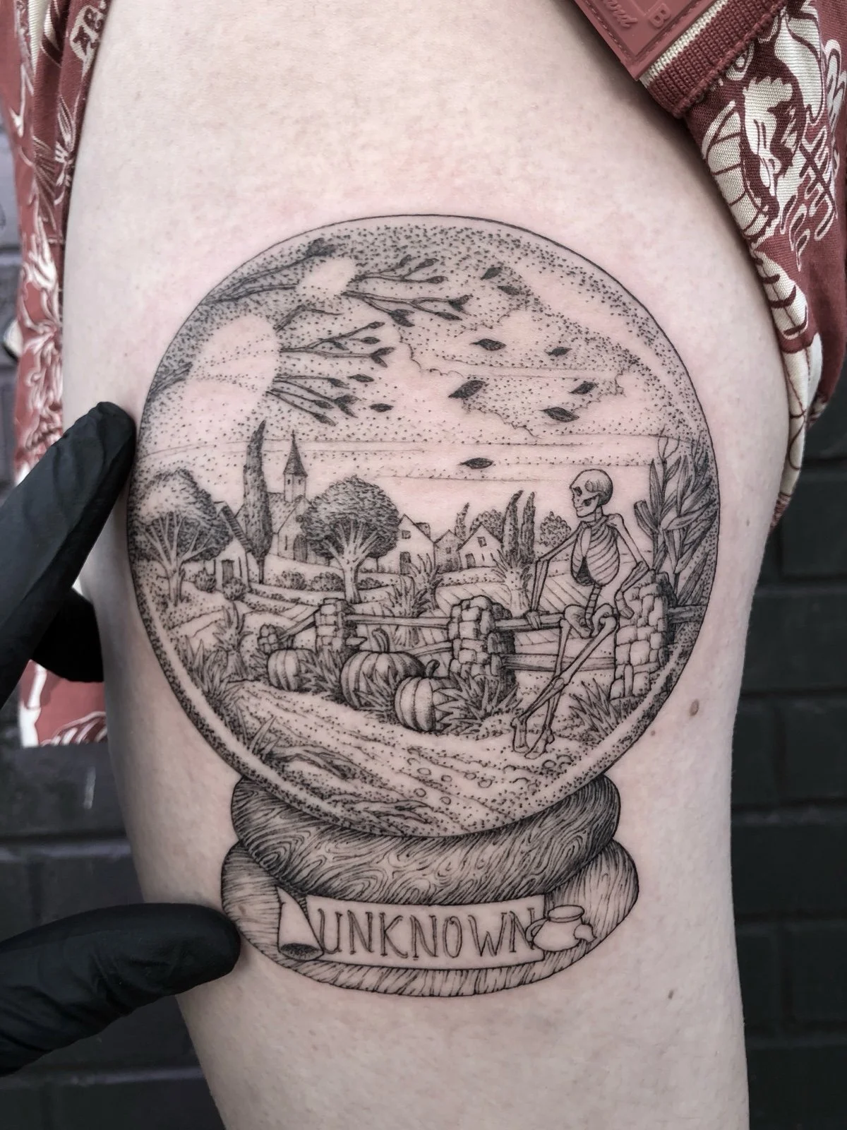 A fine line and black and grey tattoo with dotwork shading, depicting a snow globe with a skeleton looking over a New England landscape, created by Seattle tattoo artist Eli Bischof.