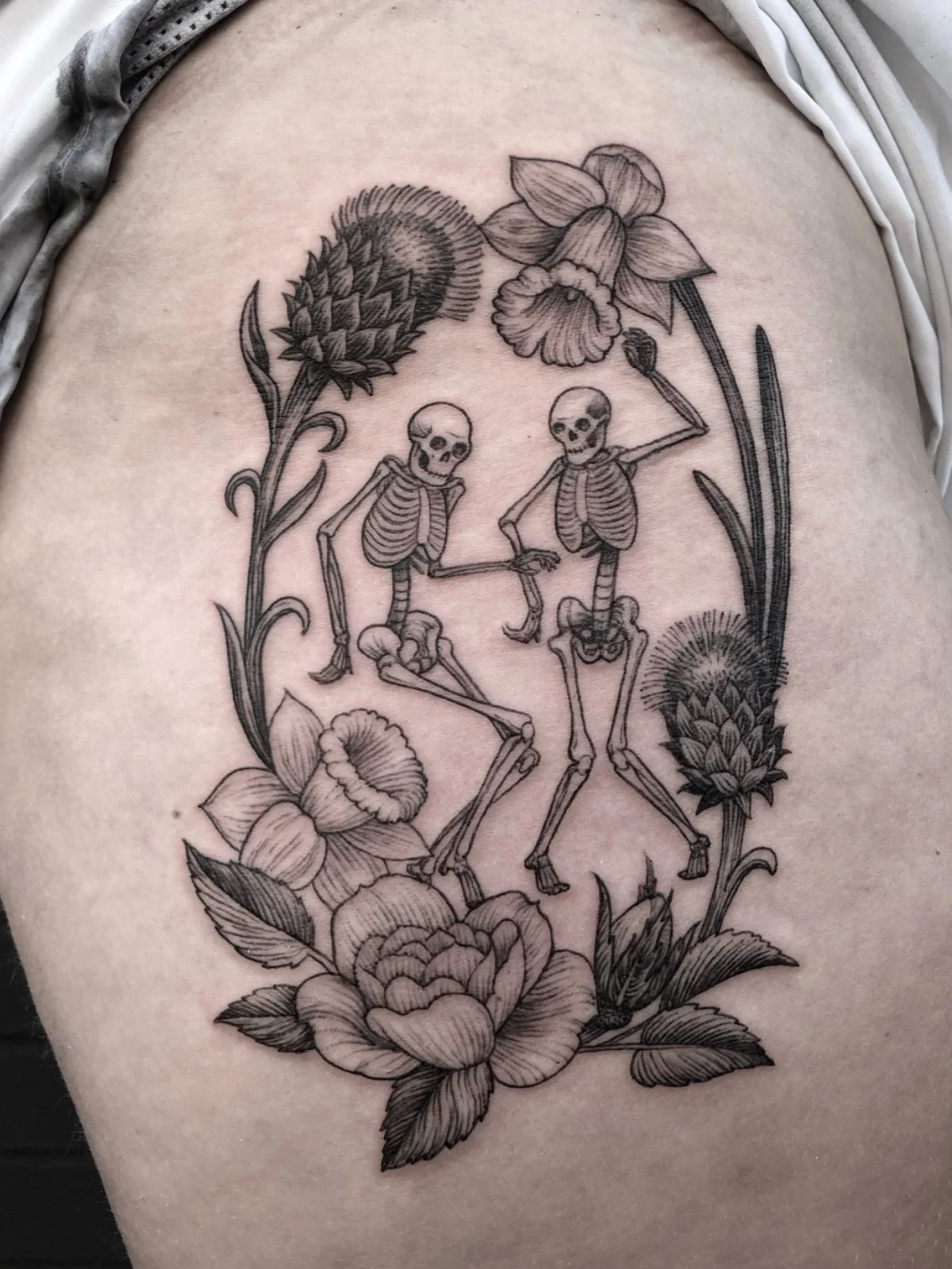Fine line black and grey thigh tattoo of dancing skeletons framed by flowers and leaves, created by Seattle tattoo artist Eli Bischof