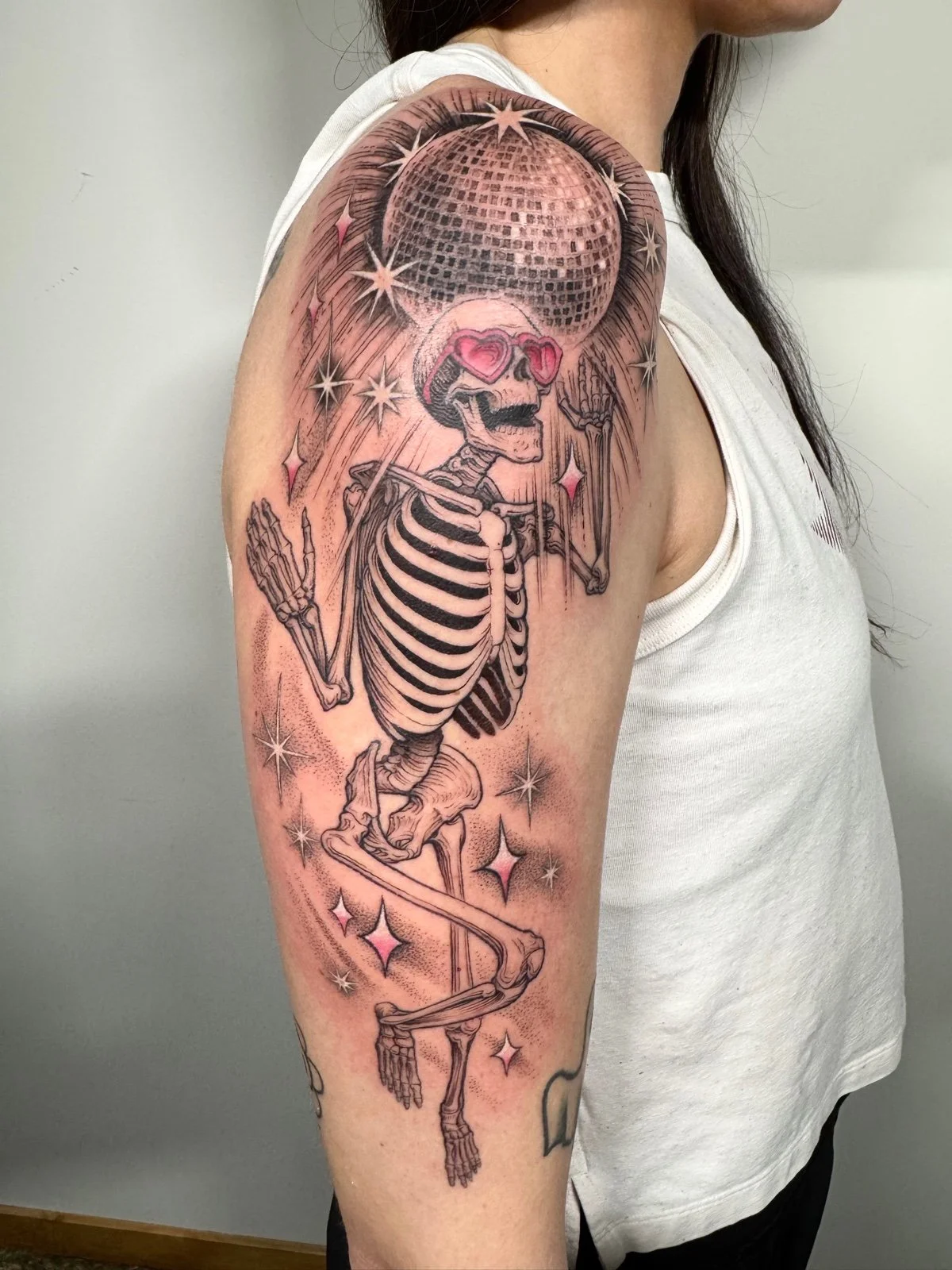 Fine line black and grey tattoo of a skeleton with pink heart shaped glasses, dancing beneath a disco ball, created by Seattle tattoo artist Eli Bischof.