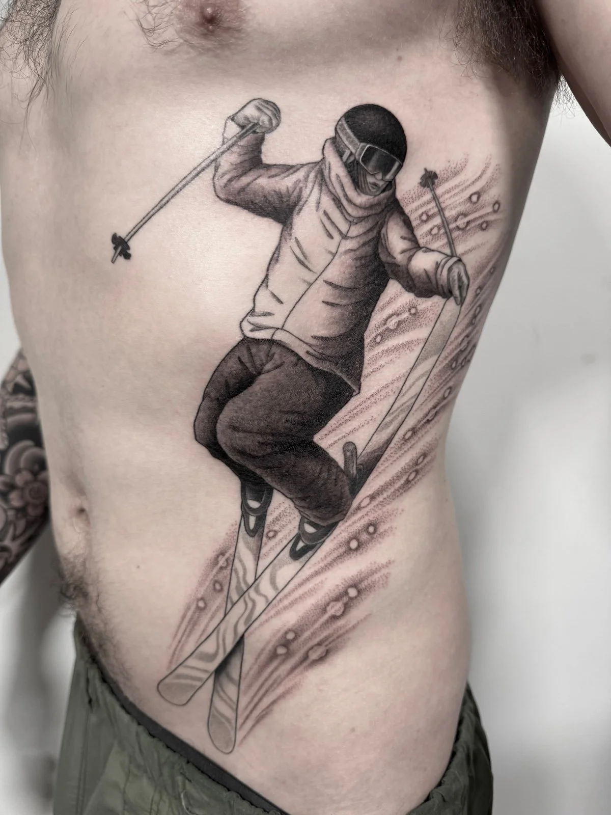 A fine line and black and grey tattoo of a skier doing a blunt grab on the torso, created by Seattle tattoo artist Eli Bischof.