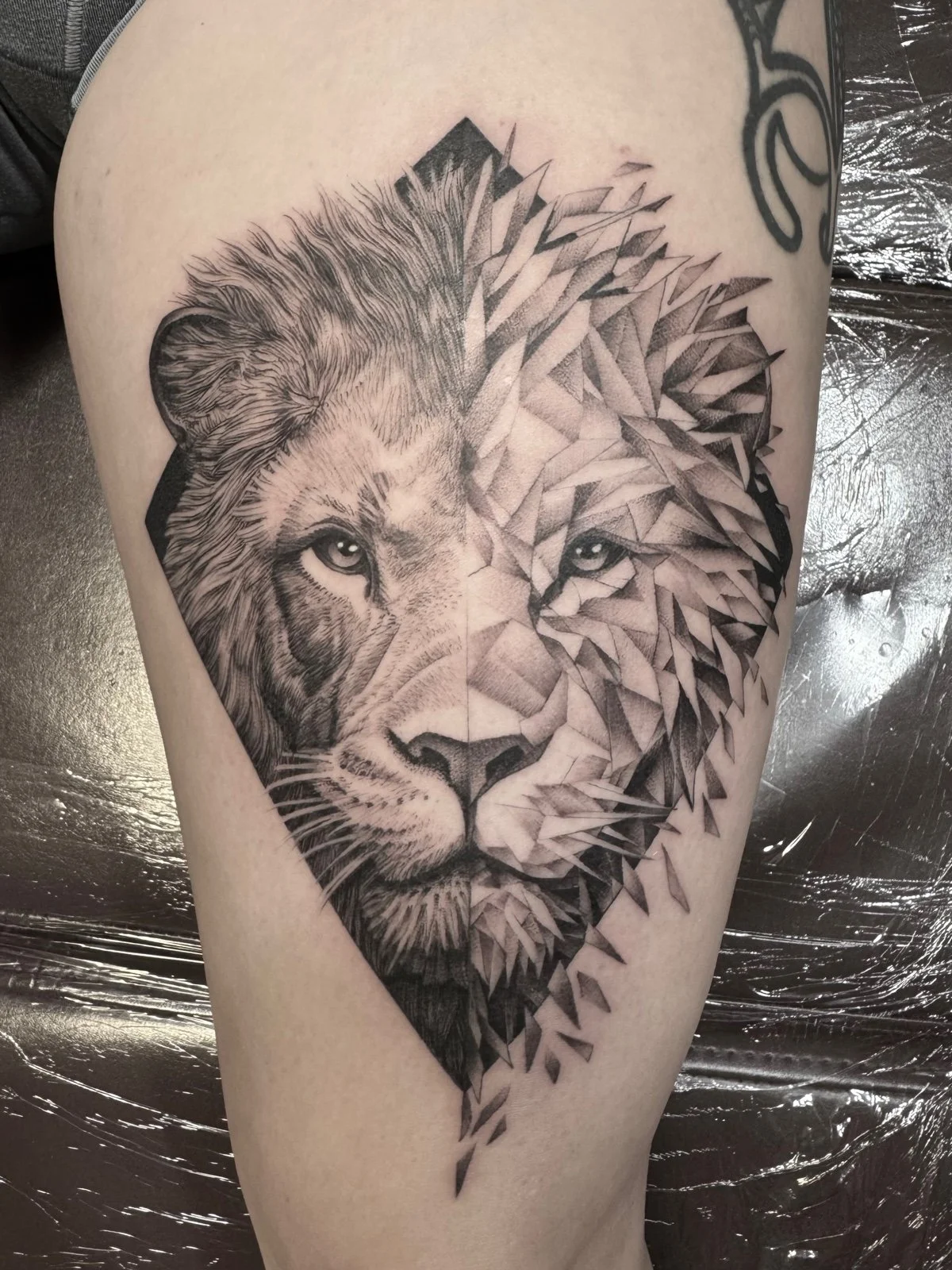 A fine line black and grey thigh tattoo of an African lion face, with half of its face rendered realistically and the other half breaking into fragments and shards, created by Seattle tattoo artist Eli Bischof.