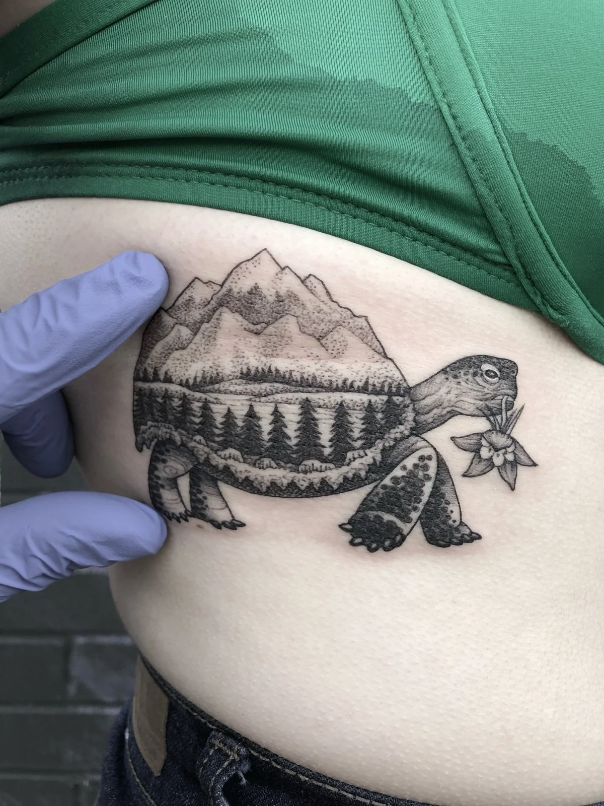 A fine line black and grey tattoo of a tortoise with a flower in its mouth, with a PNW landscape within its shell, created by Seattle tattoo artist Eli Bischof.