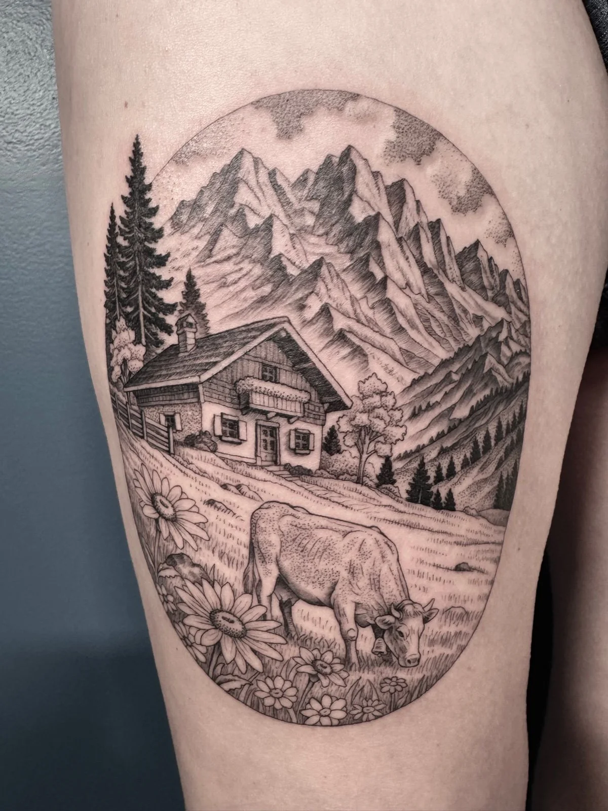 A fine line black and grey thigh landscape tattoo, featuring a cabin in the Swiss Alps and a cow grazing in a flowery meadow, created by Seattle tattoo artist Eli Bischof.