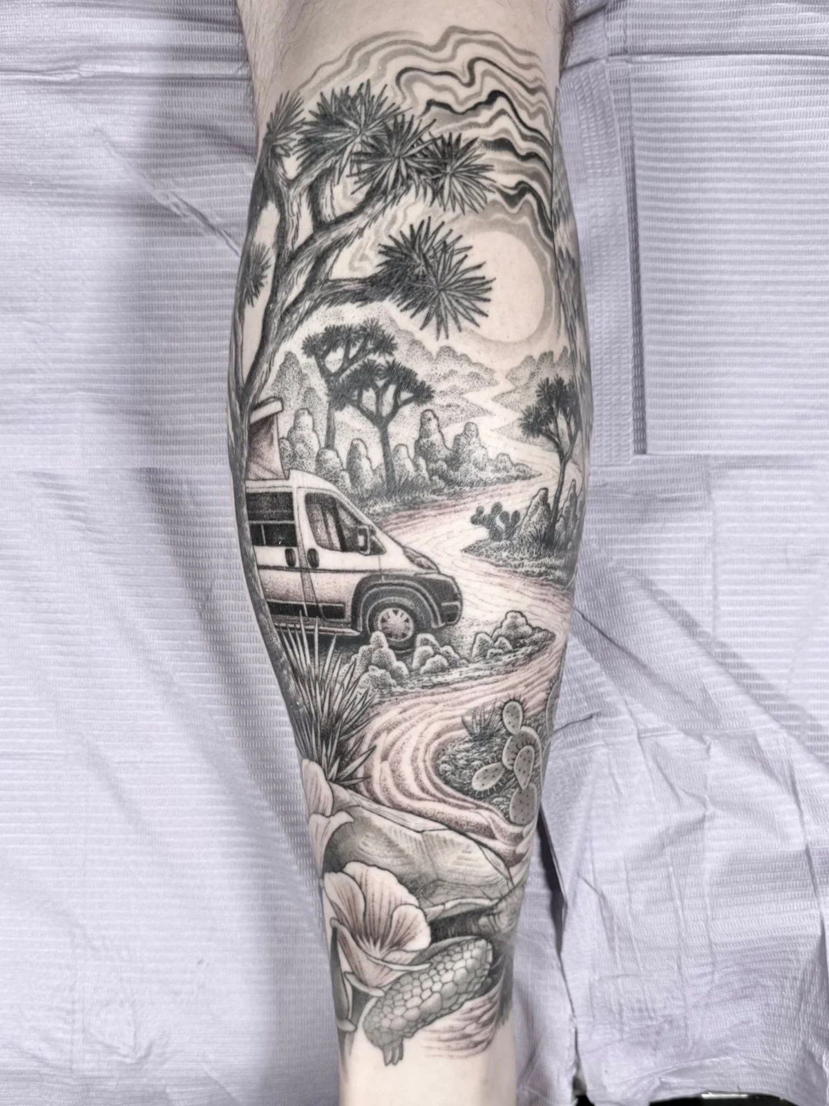 A fine line black and grey tattoo with a road winding through Joshua Tree national park and a camper van, created by Seattle tattoo artist Eli Bischof.
