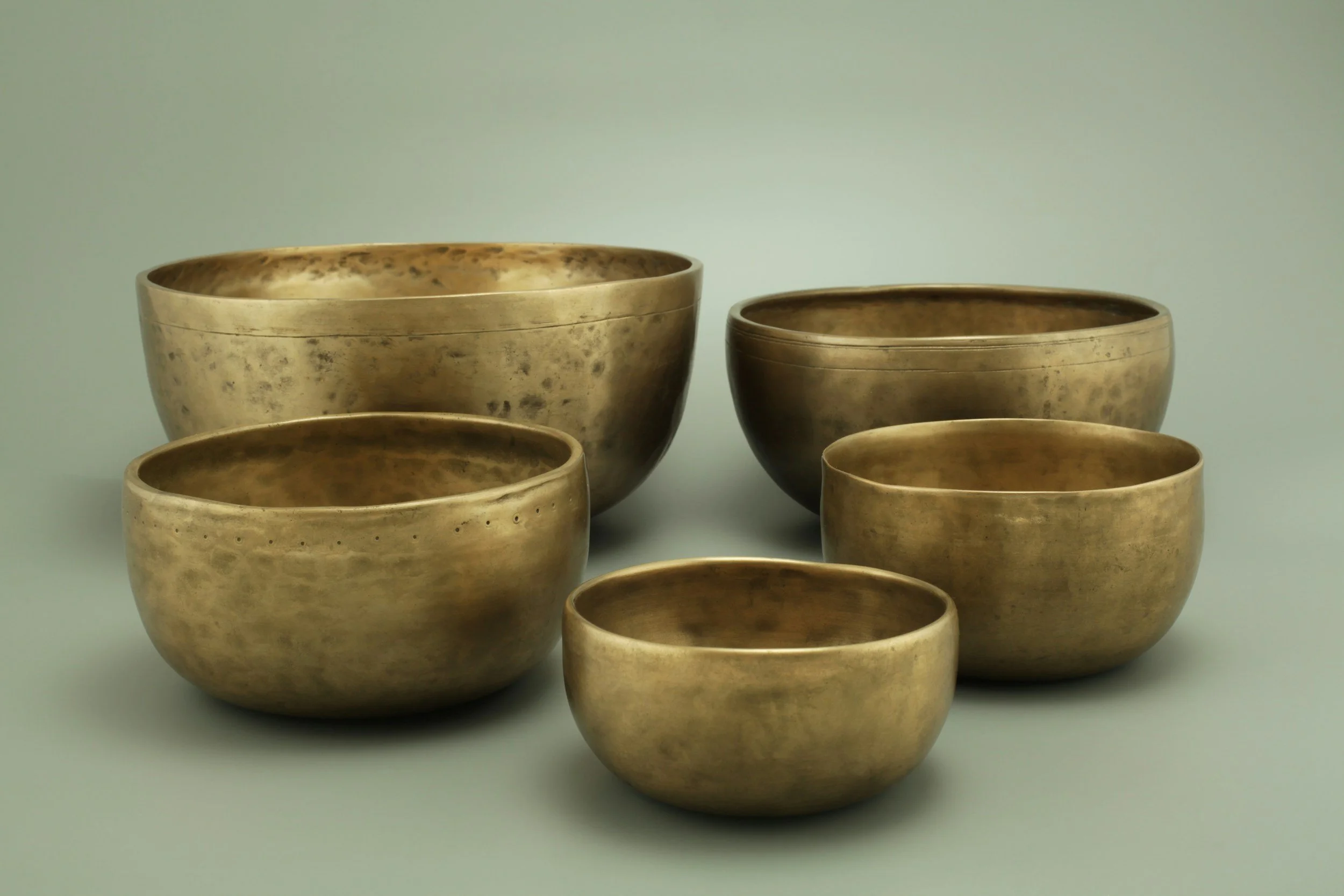 A collection of seven brass or bronze bowls of varying sizes arranged against a plain light-colored background.