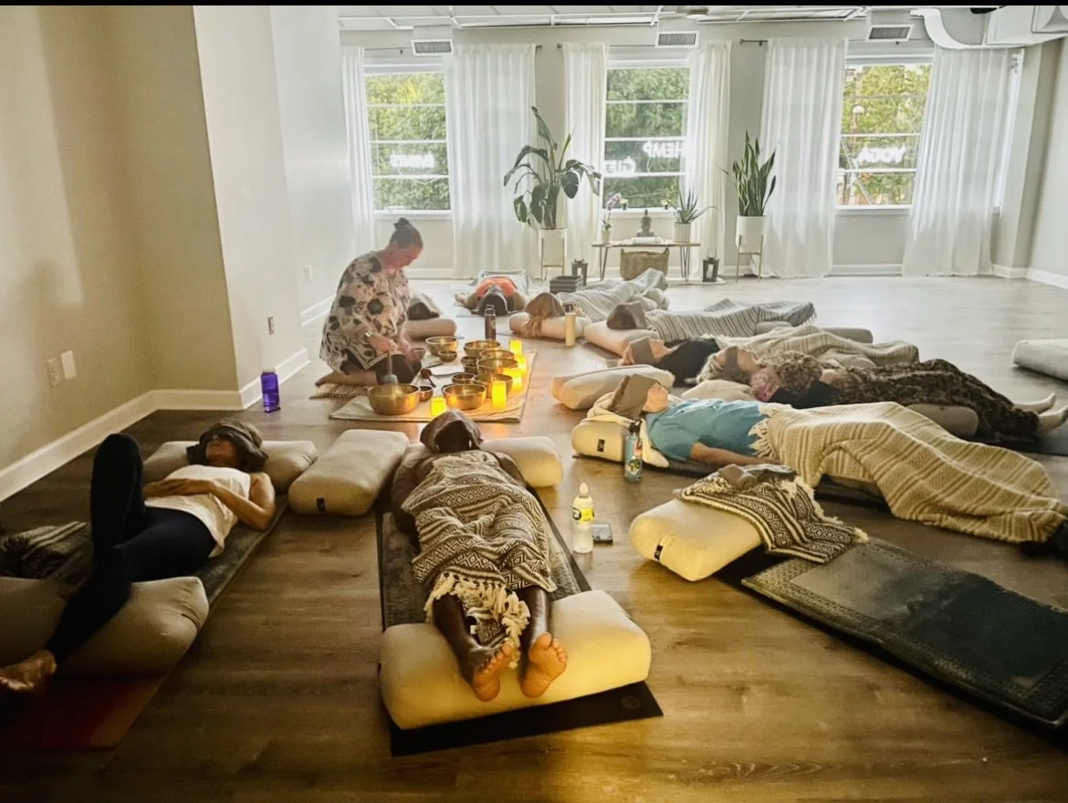 People lying on mats and cushions on the floor of a spacious, dim-lit room with large windows and white curtains. A person is sitting cross-legged in front of a row of lit candles and bowls, playing Tibetan singing bowls
