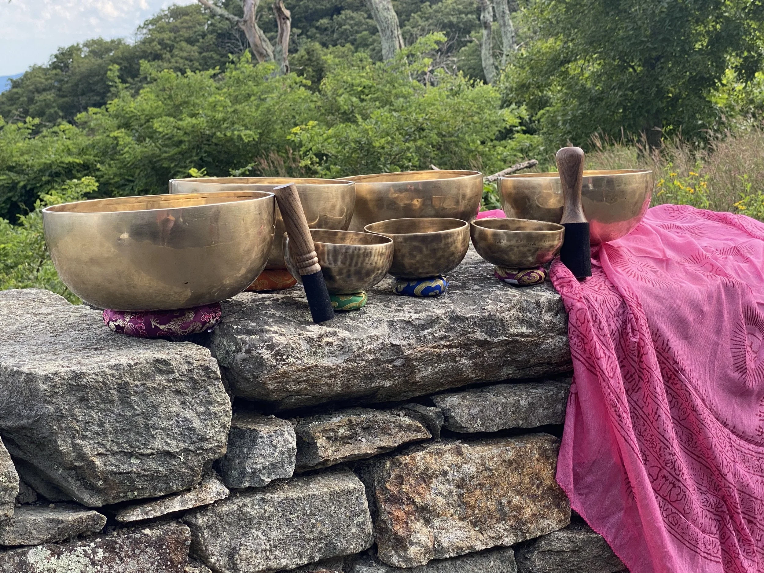 Several singing bowls of different sizes placed on a rock wall with a colorful Tibetan cloth underneath, set in an outdoor natural environment with trees and greenery in the background.