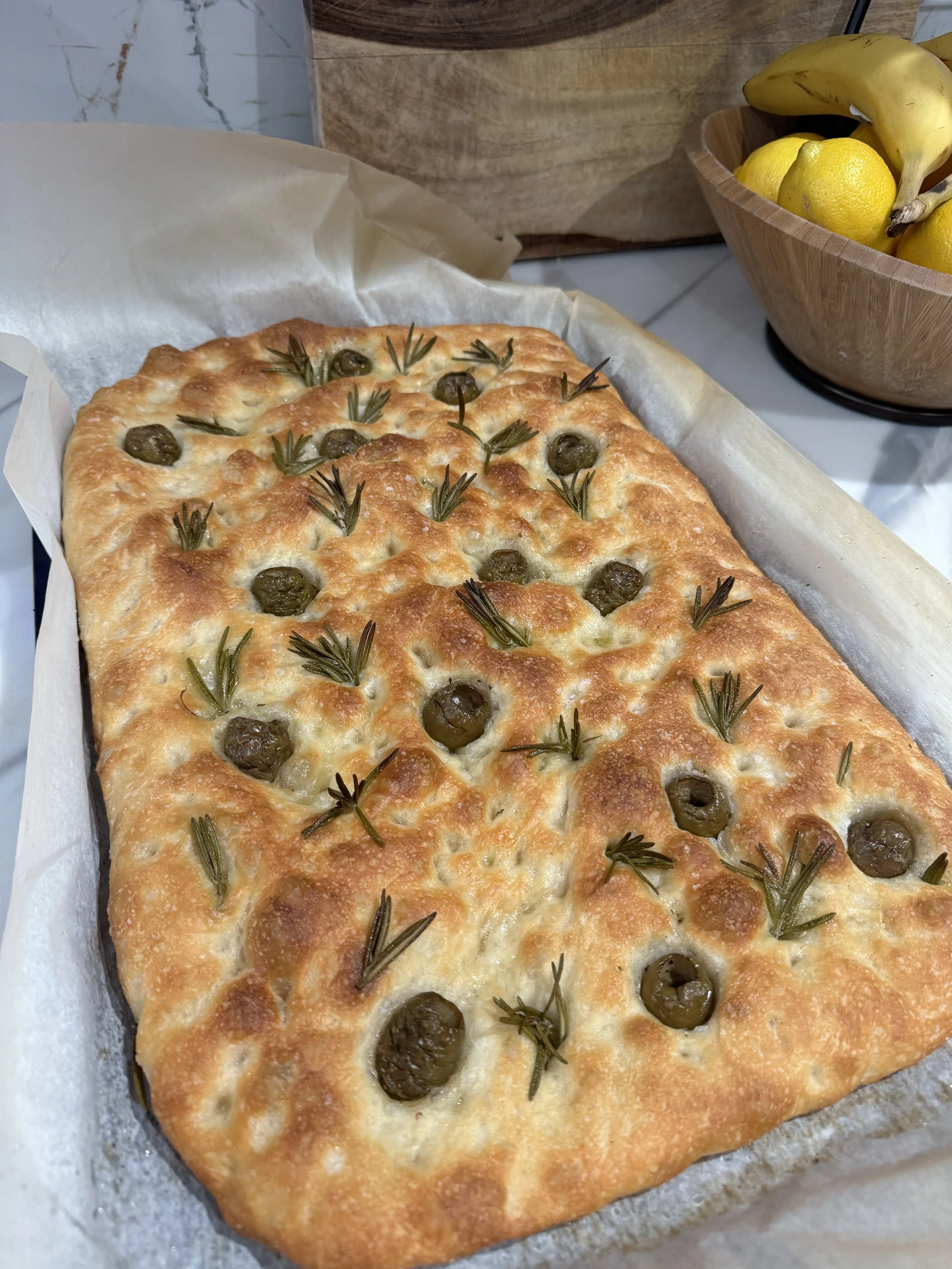 Baked flatbread topped with green olives and fresh rosemary, placed on parchment paper.