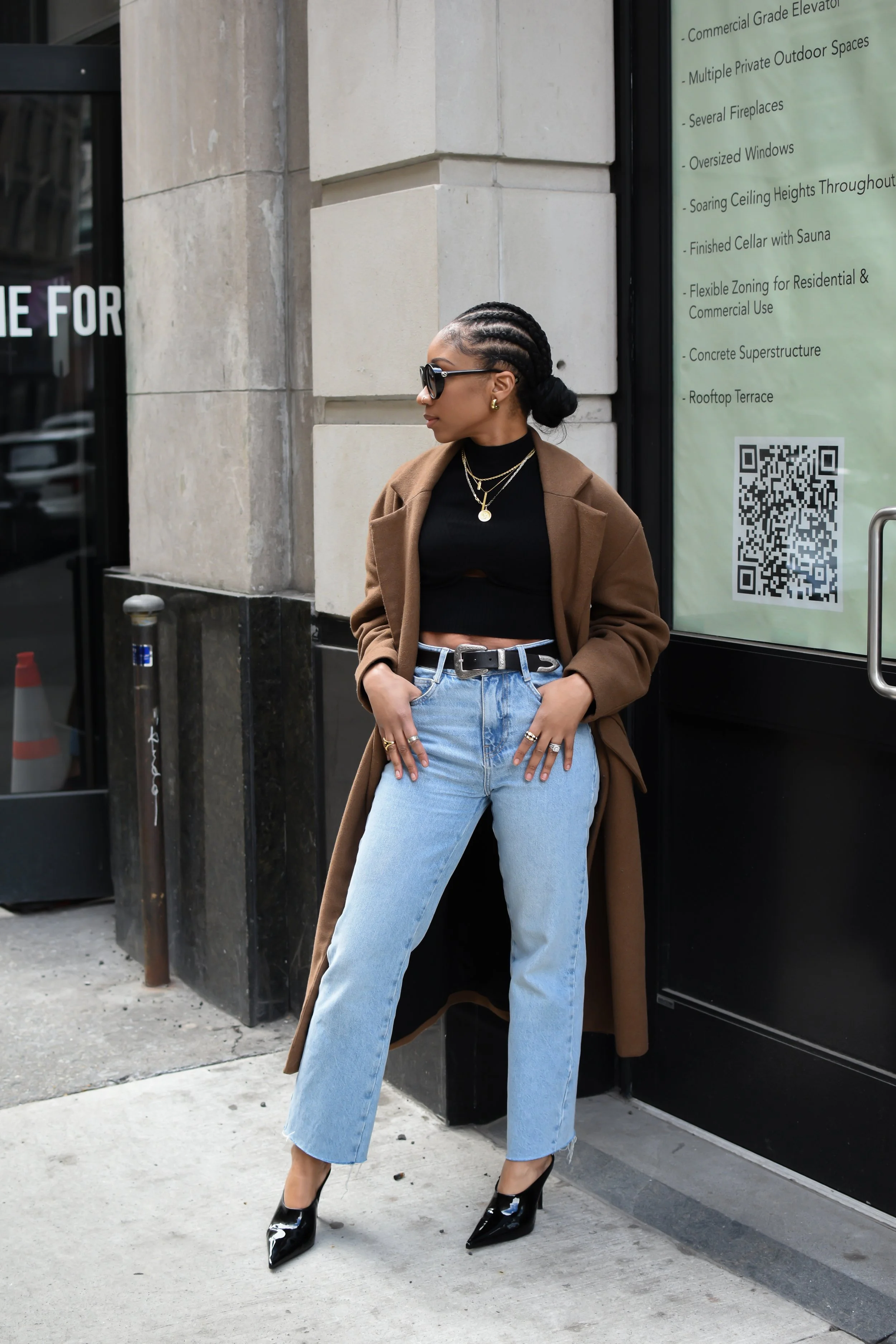 A woman standing on city sidewalk wearing sunglasses, a black crop top, light blue jeans, black heels, and a brown coat, with her hands in her pockets, near a building with stone walls and a large window with a sign.
