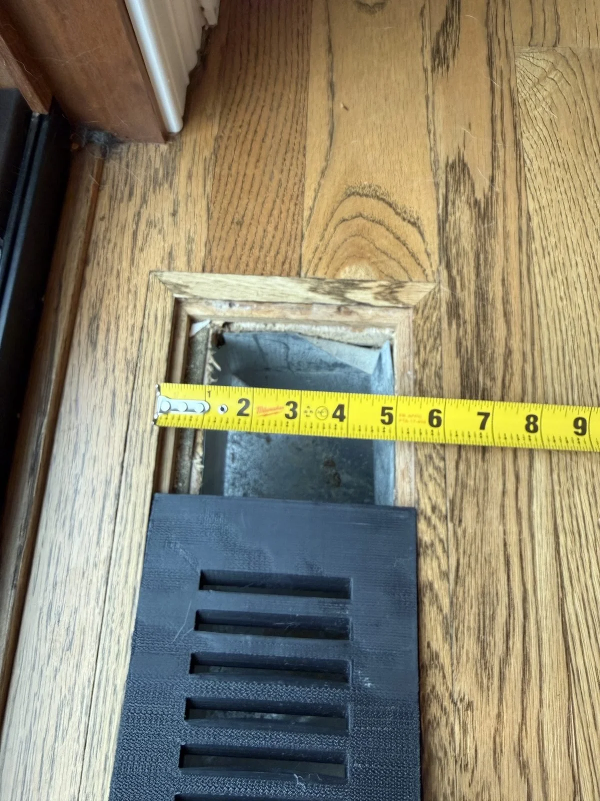 Tape measure across the width of a recessed floor vent opening in a hardwood floor