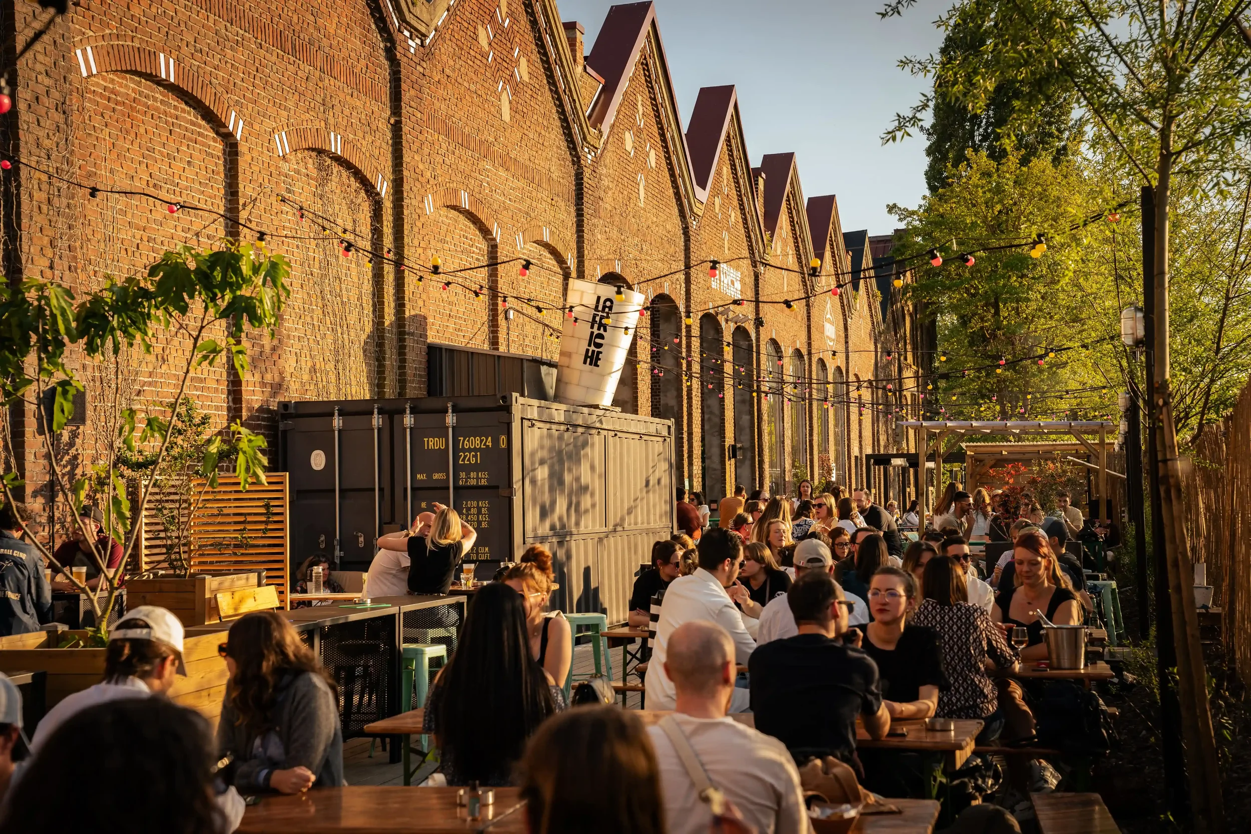 Un espace extérieur d'un restaurant ou bar animé avec de nombreux clients assis à des tables en bois, entouré de murs en briques et de luminaires décoratifs suspendus. L'ambiance est chaleureuse, avec de la végétation et des arbres, sous un ciel clai