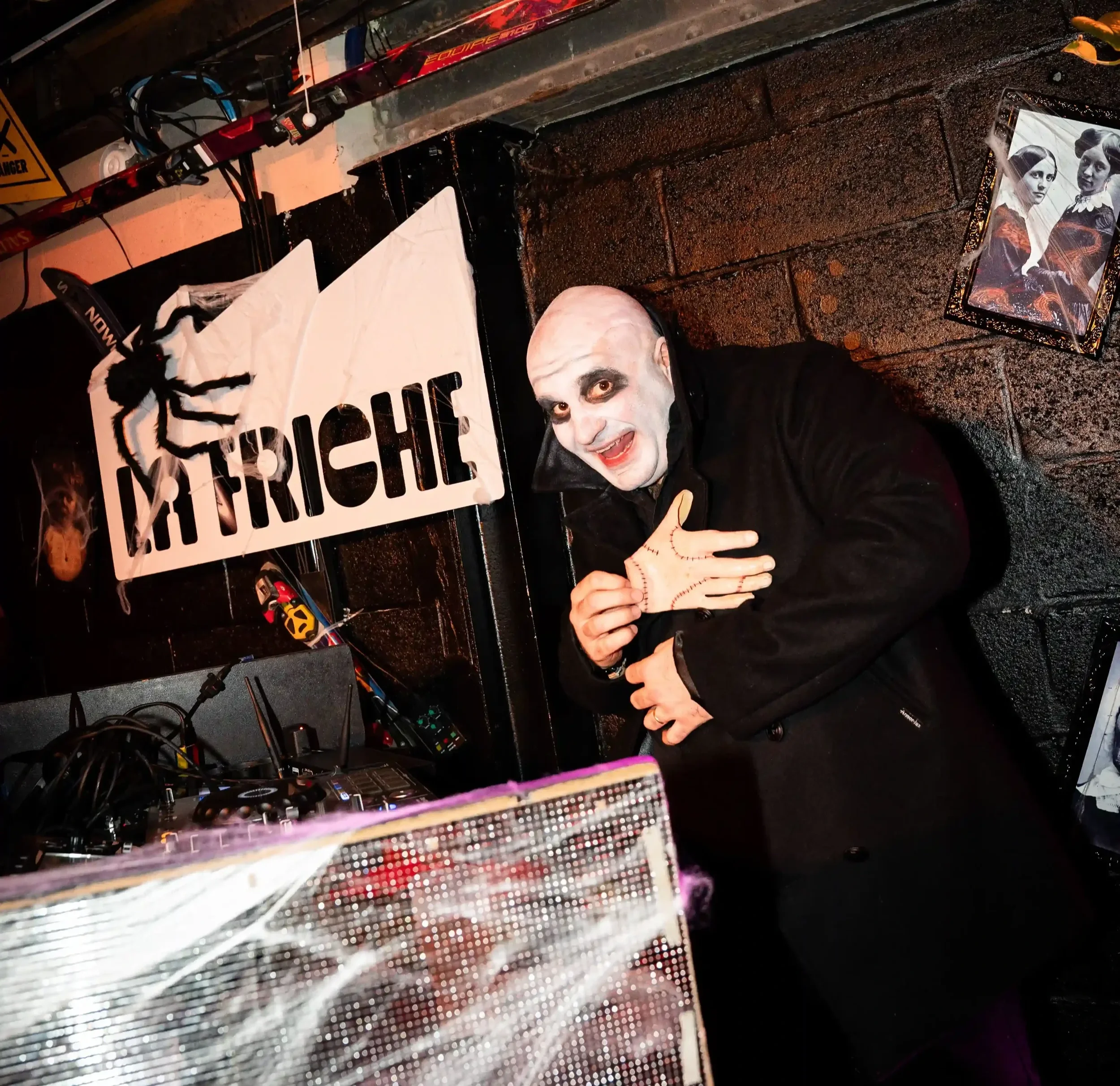 Un homme déguisé en clown avec un visage peint en blanc, portant un manteau noir, sourit et fait un geste avec sa main. Derrière lui, il y a une affiche avec une araignéefact, une maison en briques noires, et une photo encadrée de deux femmes en robes anciennes. La scène semble être dans un décor de fête ou de spectacle.